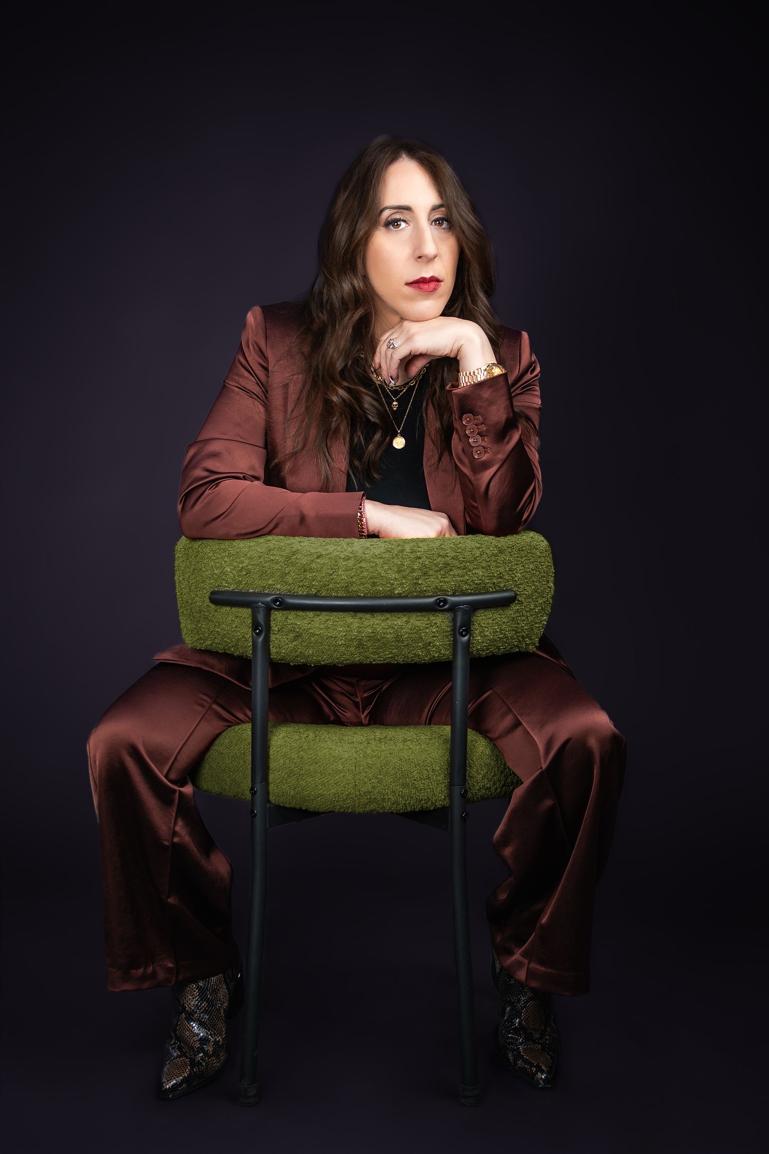 A person in a brown suit sits backwards on a green chair against a dark background, striking a contemplative pose.
