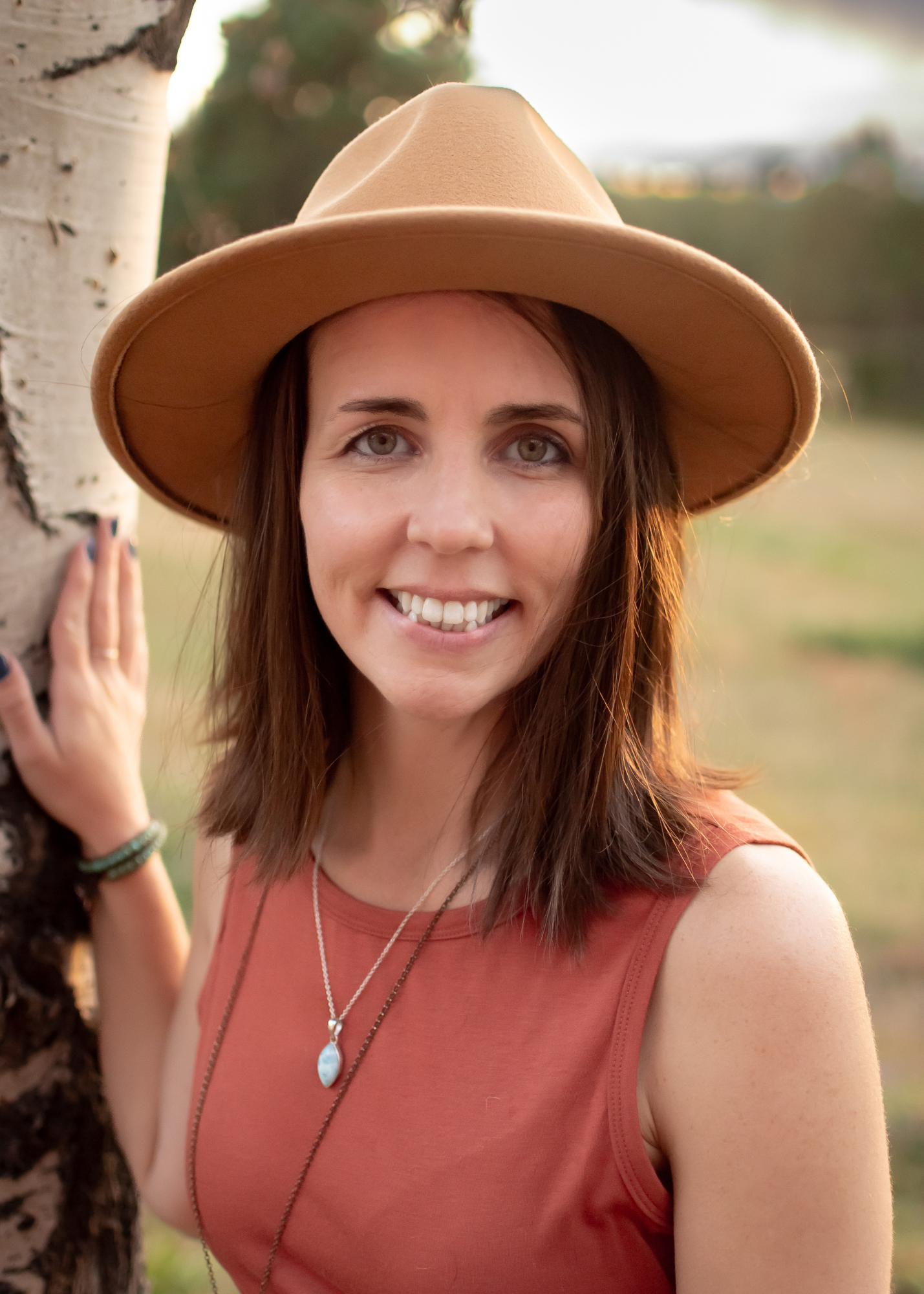 Woman in brown hat and sleeveless rust-colored top poses outdoors in warm sunlight.