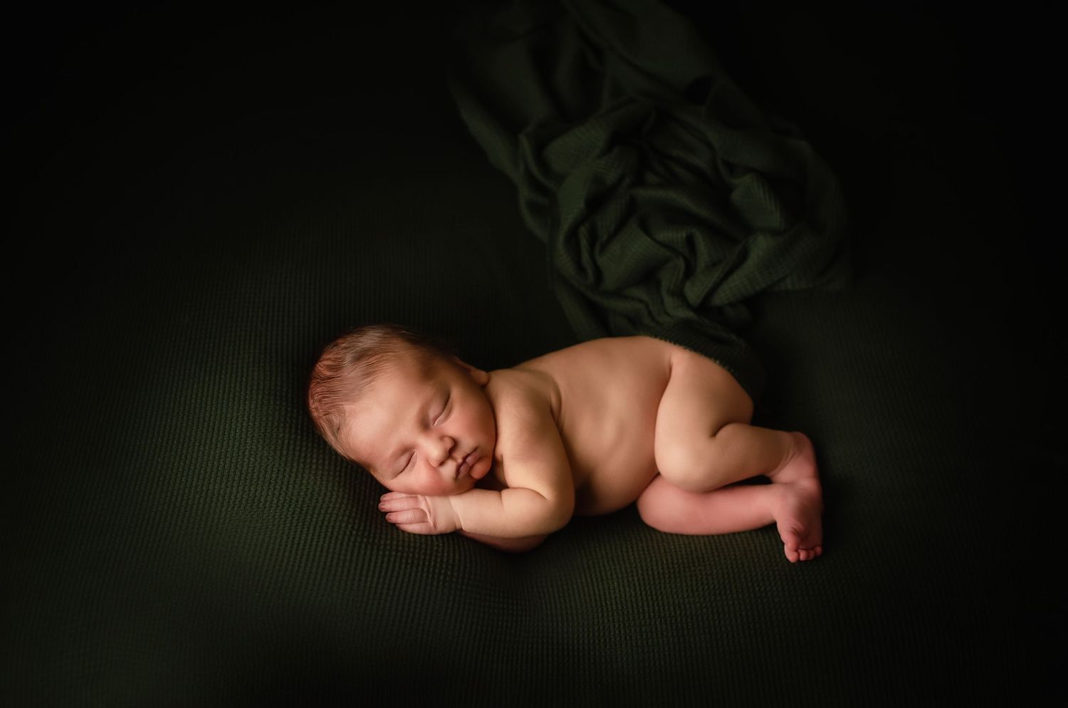Sleeping newborn baby posed on dark green backdrop in artistic lighting.