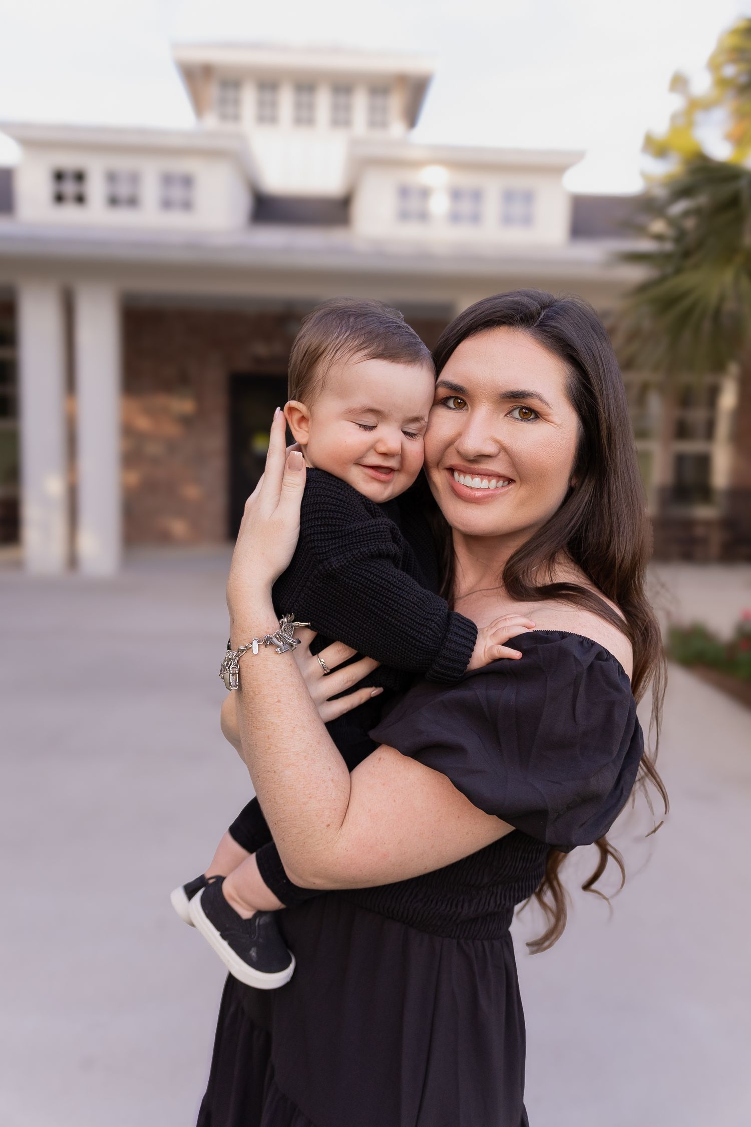Smiling dark-haired woman in black dress holding a baby boy outdoors.