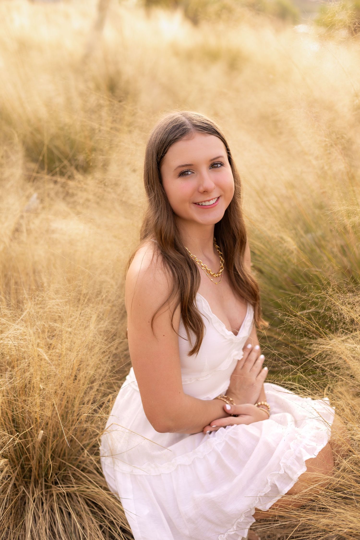 Smiling teenage girl in white dress sitting in golden tall grass field during sunset portrait.