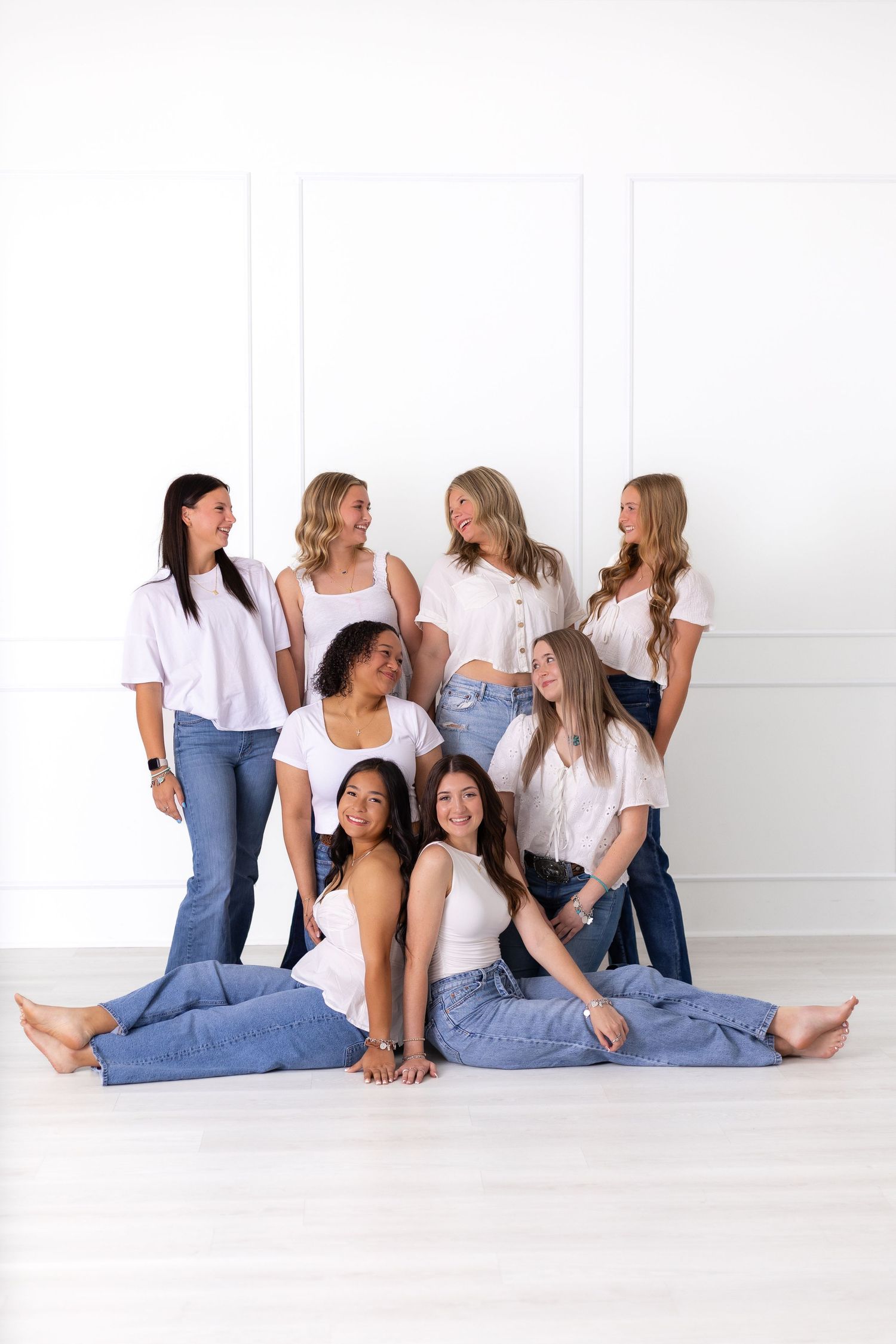 Seven diverse women in white tops and jeans posing together in a bright white studio setting.