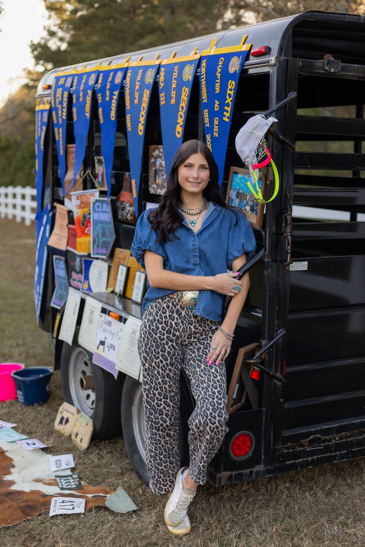Smiling brunette woman in denim top and leopard print pants poses beside a decorated horse trailer outdoors.