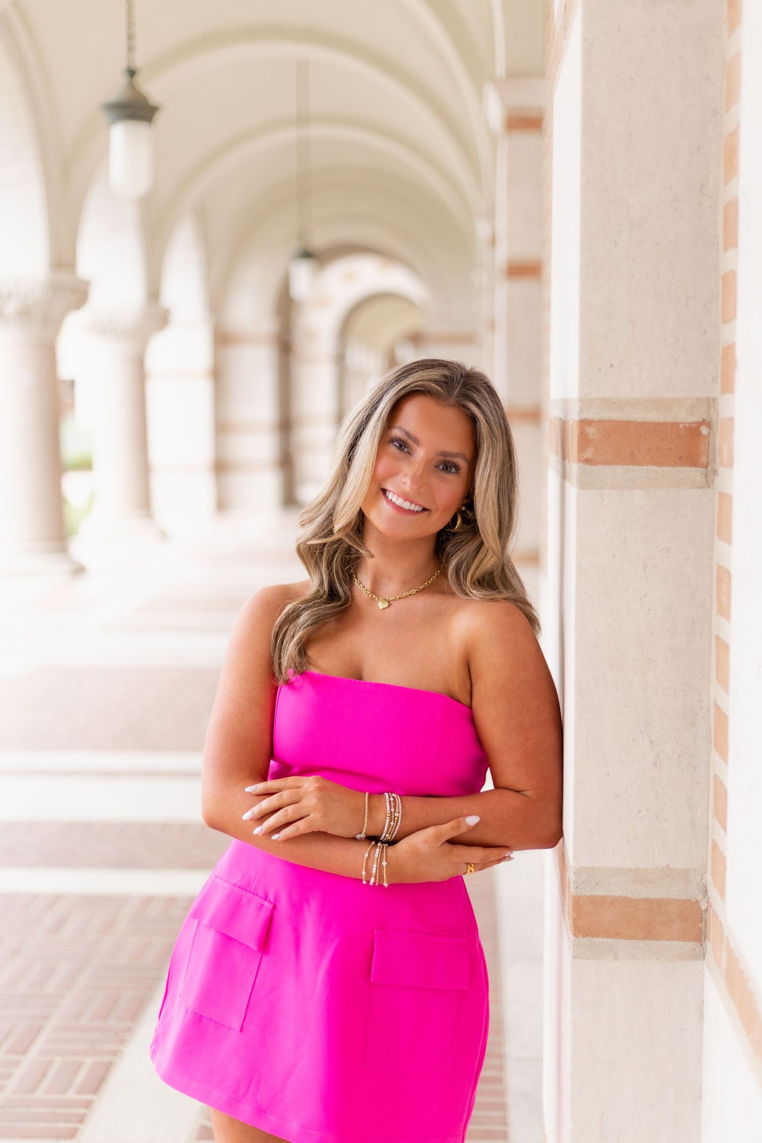 Blonde woman in hot pink strapless mini dress leans against a column in an elegant arched corridor.