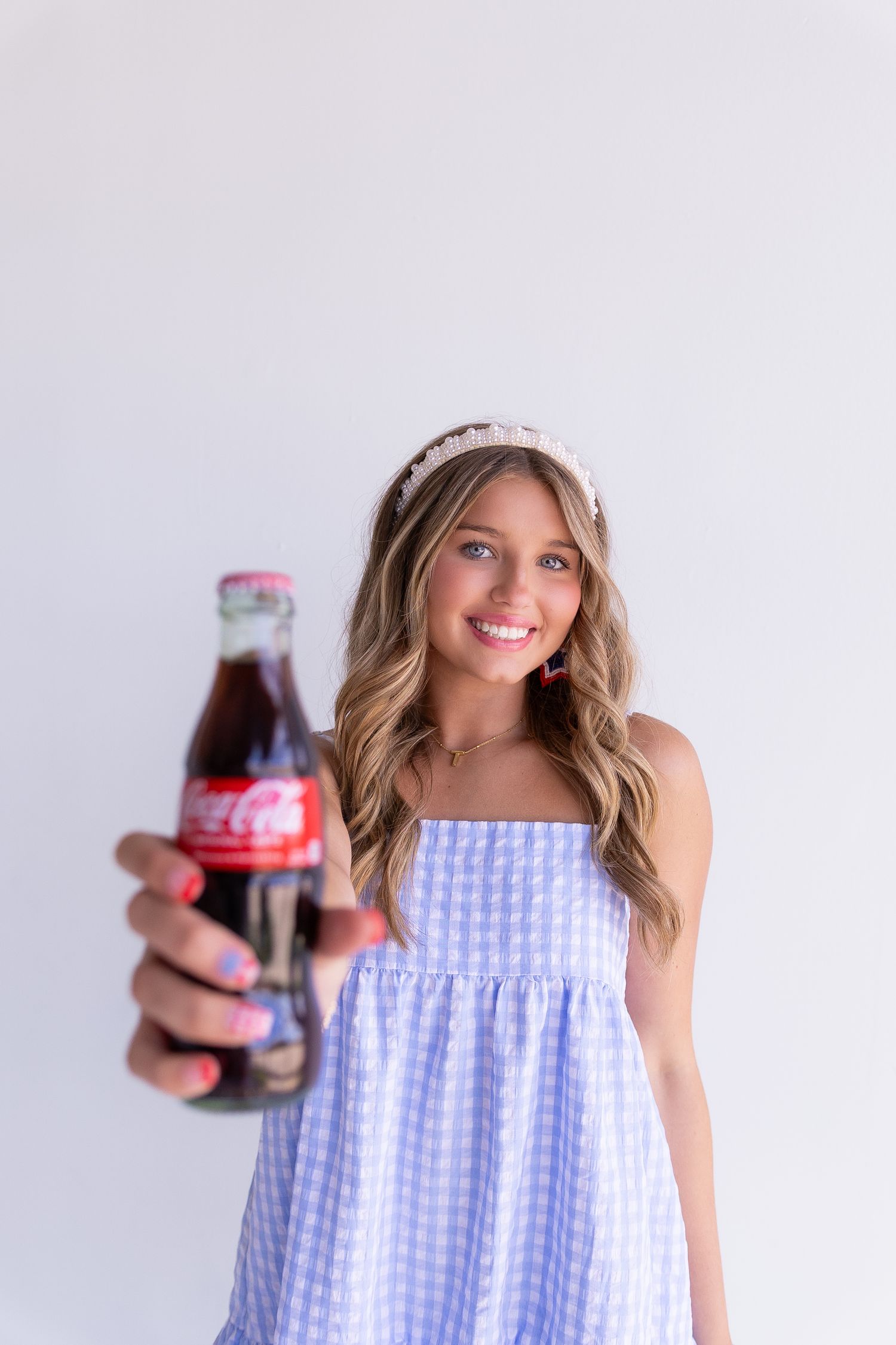 Smiling girl in blue gingham dress holds a classic Coca-Cola glass bottle toward the camera.