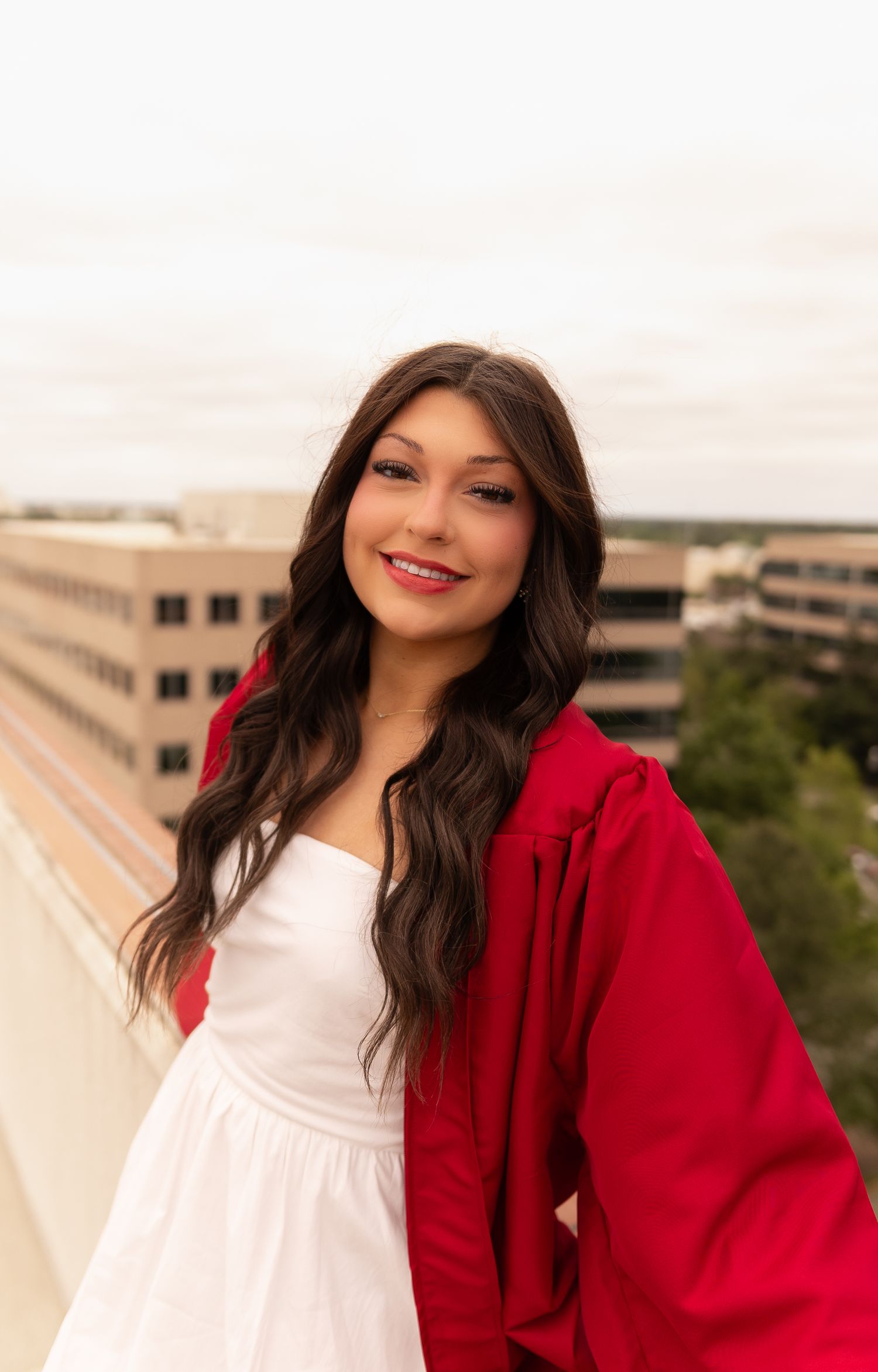 Smiling young woman in red graduation gown and white dress posing on rooftop with city view.