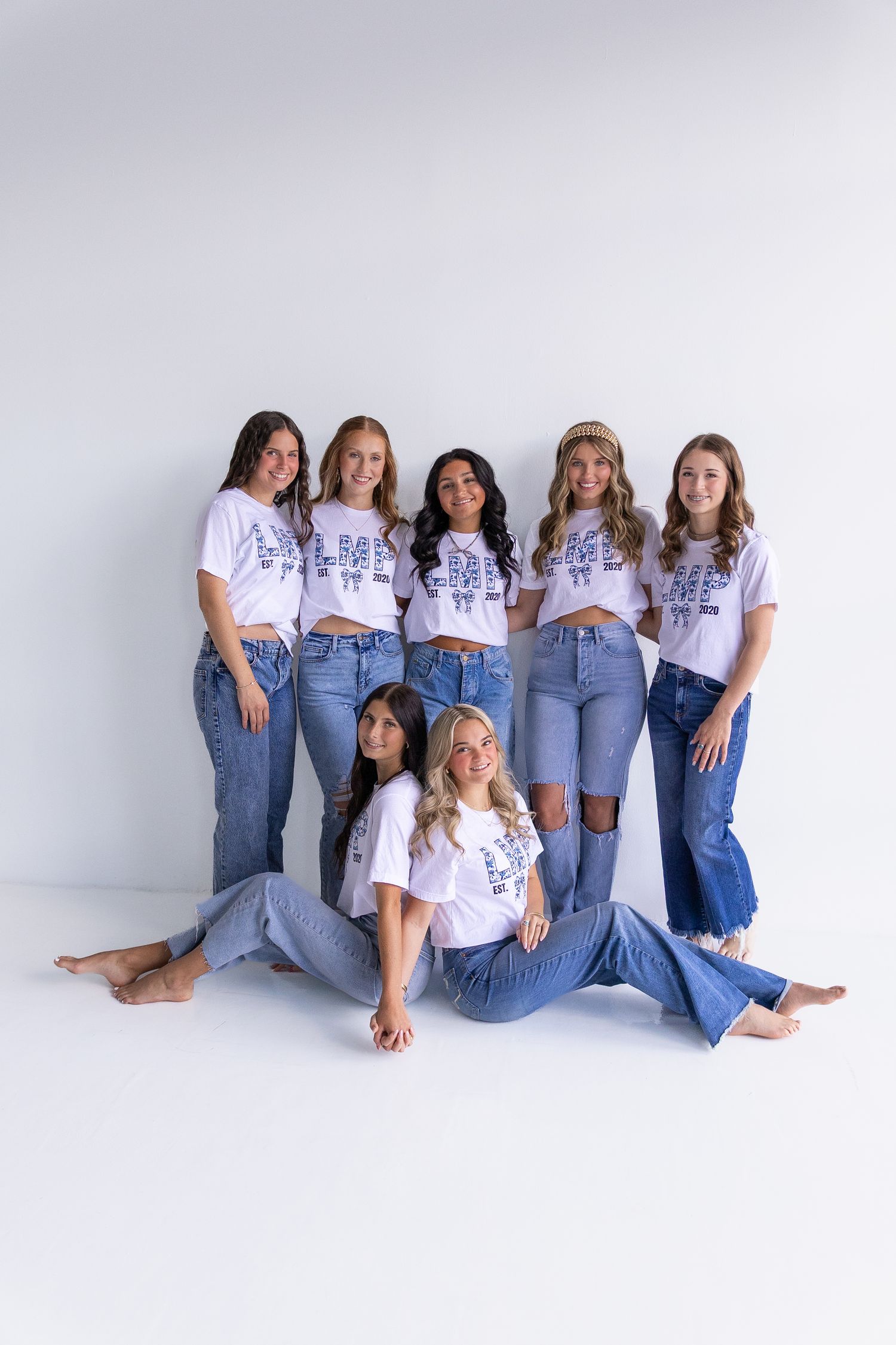 Group of seven young women in matching white graphic tees and blue jeans posing in white studio.