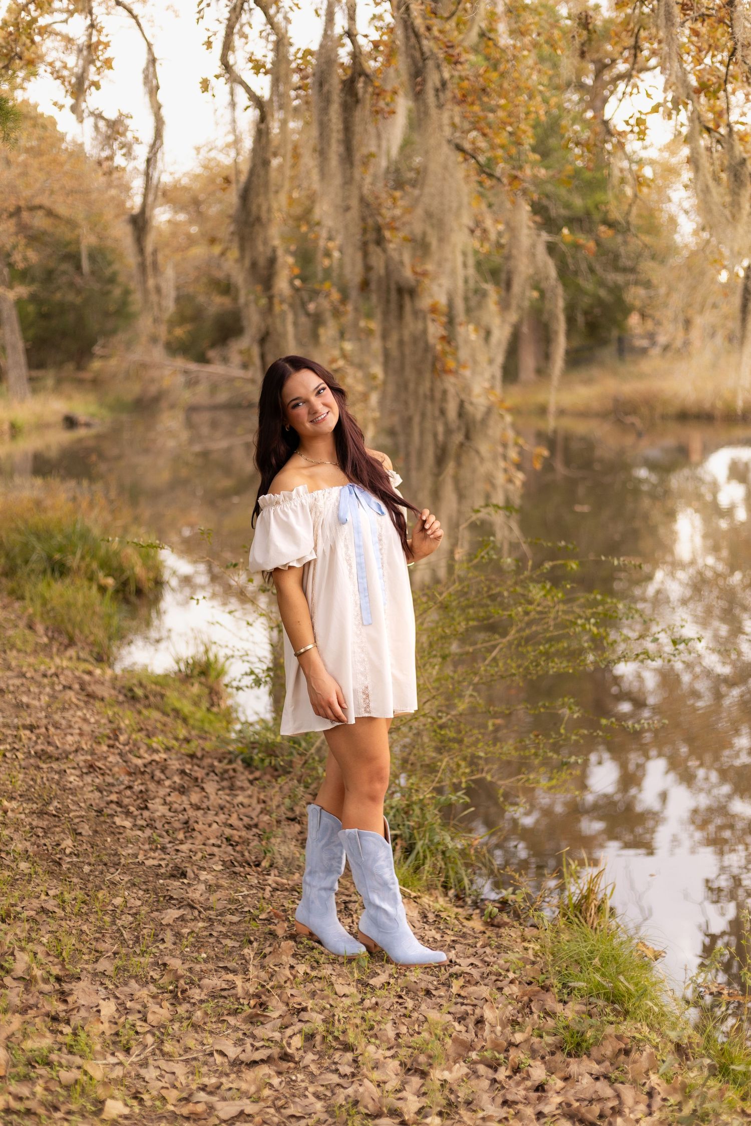 Smiling young woman in white off-shoulder dress and blue cowboy boots stands by a reflective autumn pond.