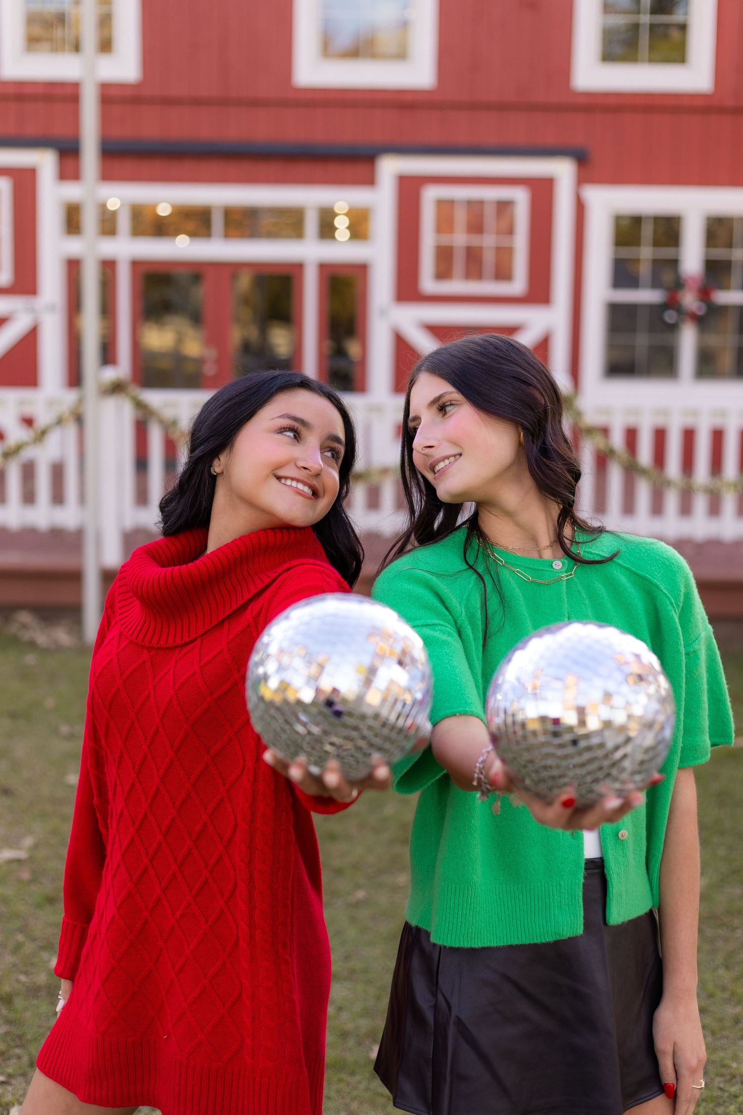 Two brunette women holding disco balls outside red barn, one in red sweater and one in green top.