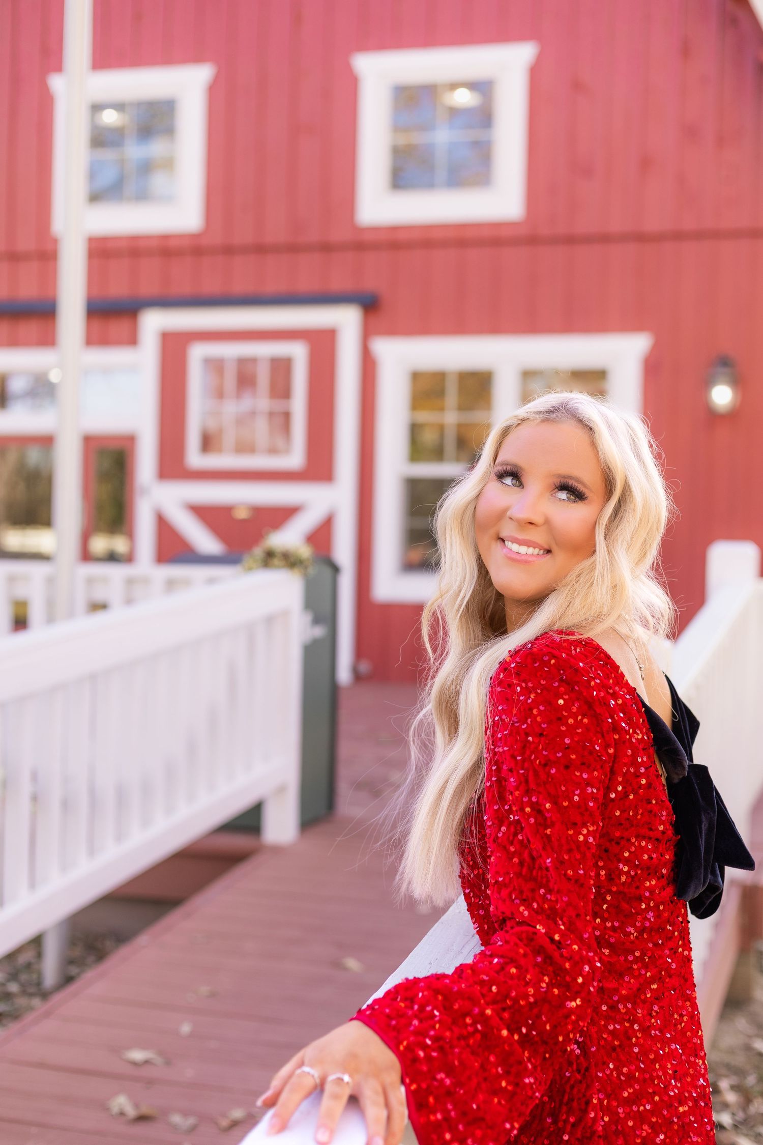 Blonde woman in red sequin jacket smiling over shoulder near white fence outside red barn.