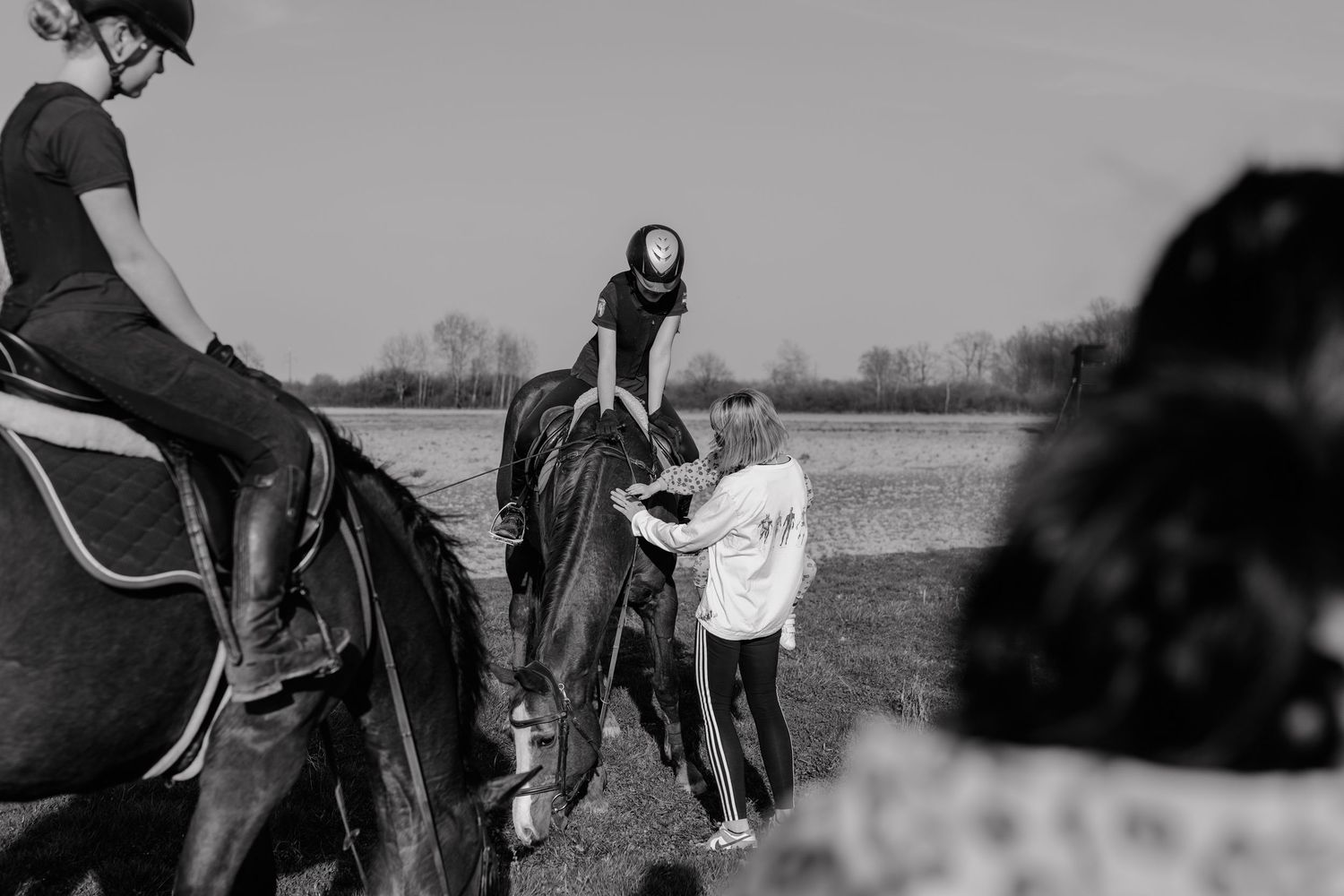 A black and white photograph shows people on horseback with others helping during a riding lesson outdoors.