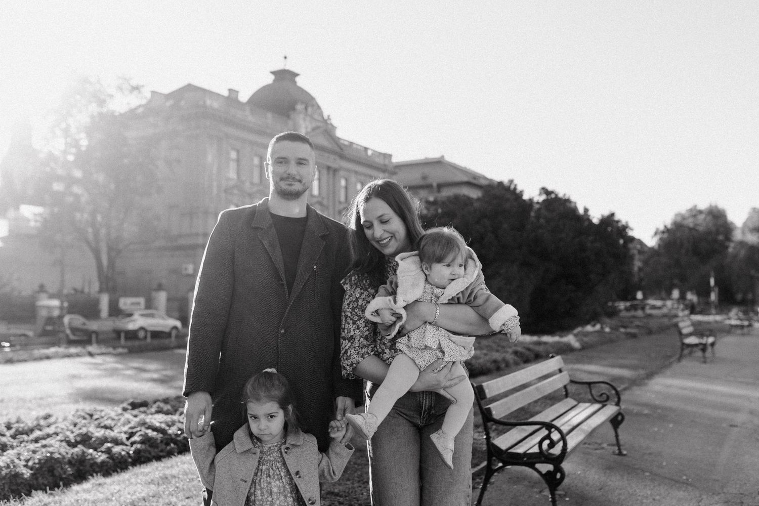 A family poses together on a sunny day in front of a historic building with park benches in the background.