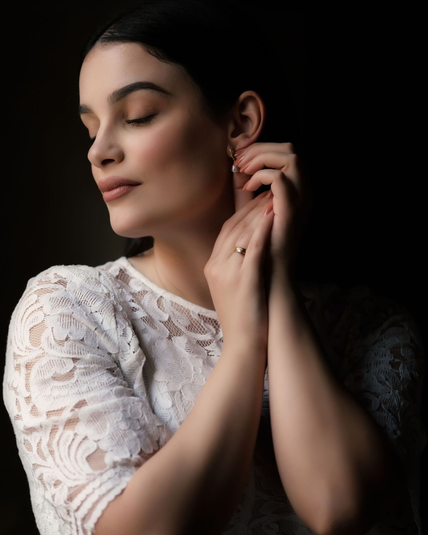 A portrait showing a serene figure in a white lace top with eyes closed and hands delicately touching against a dark background.