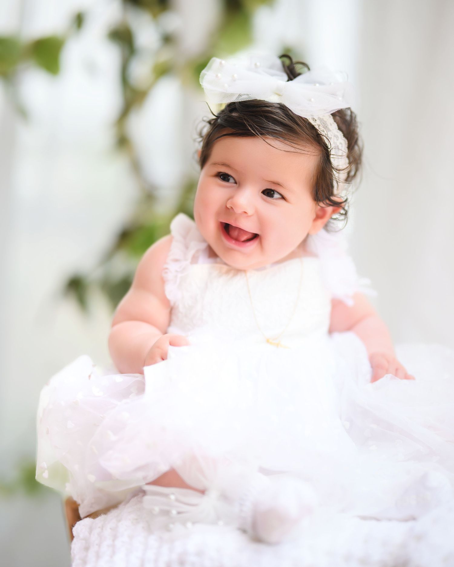 Baby wearing white dress and headband laughs joyfully while sitting against light background.