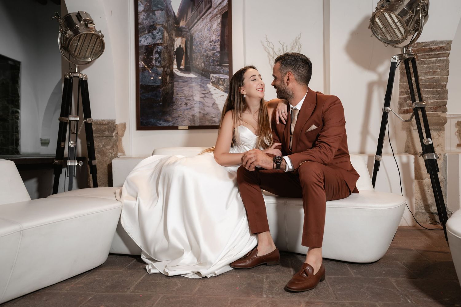 A couple relaxes on modern white chairs in an industrial-style photo studio setting.