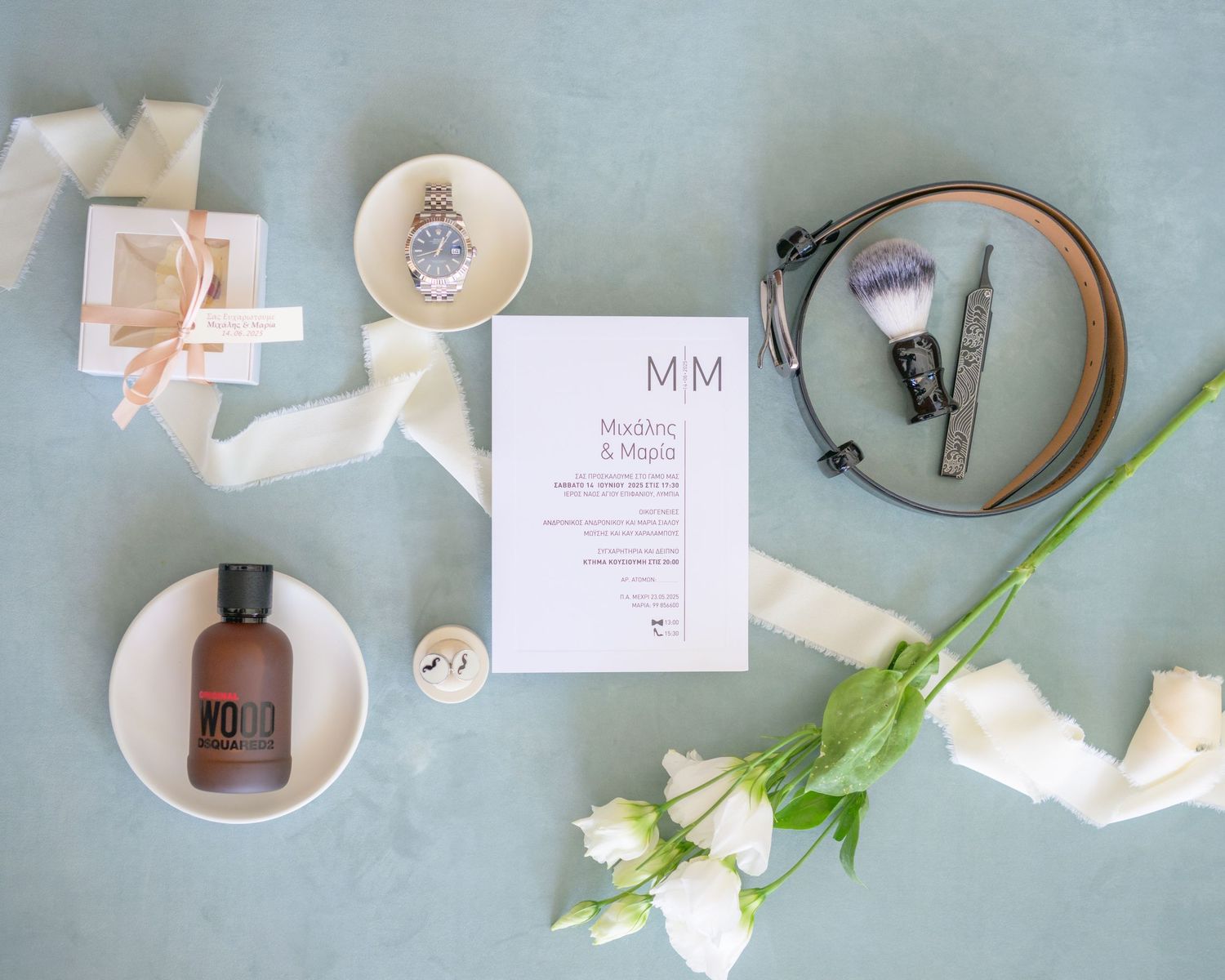 Flatlay of wedding details including white roses, ribbon, invitation card, and perfume bottle on sage green background.