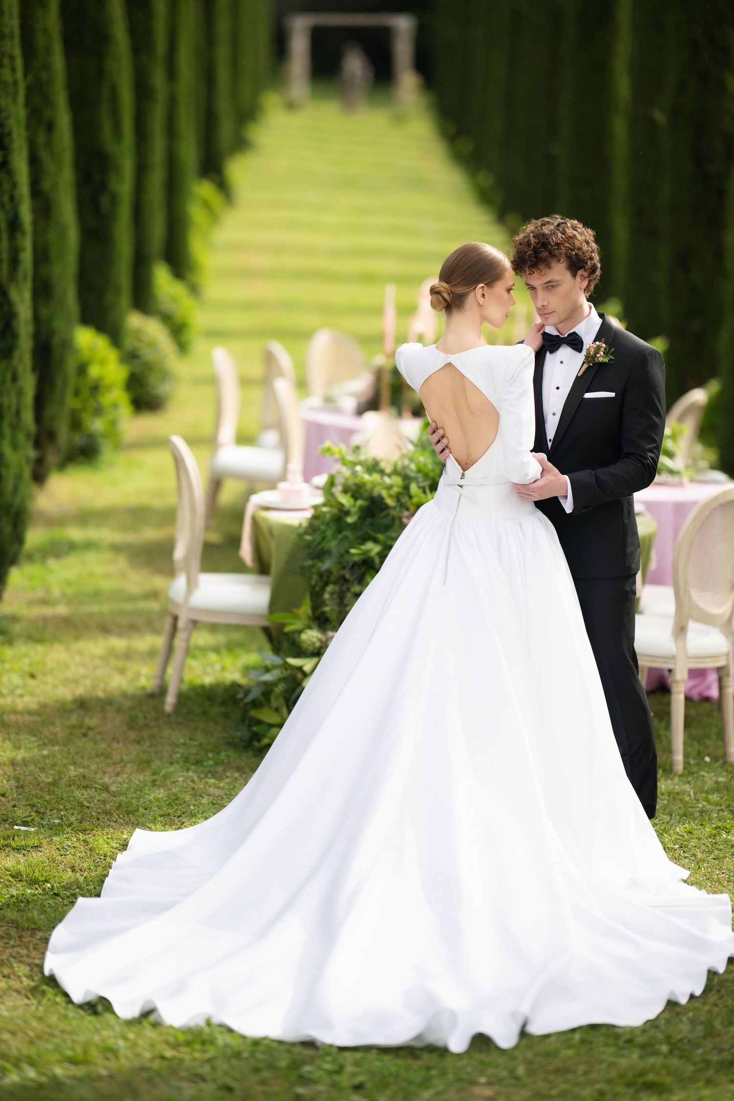 A romantic wedding couple shares intimate moments in a garden setting with cypress trees and white chairs.