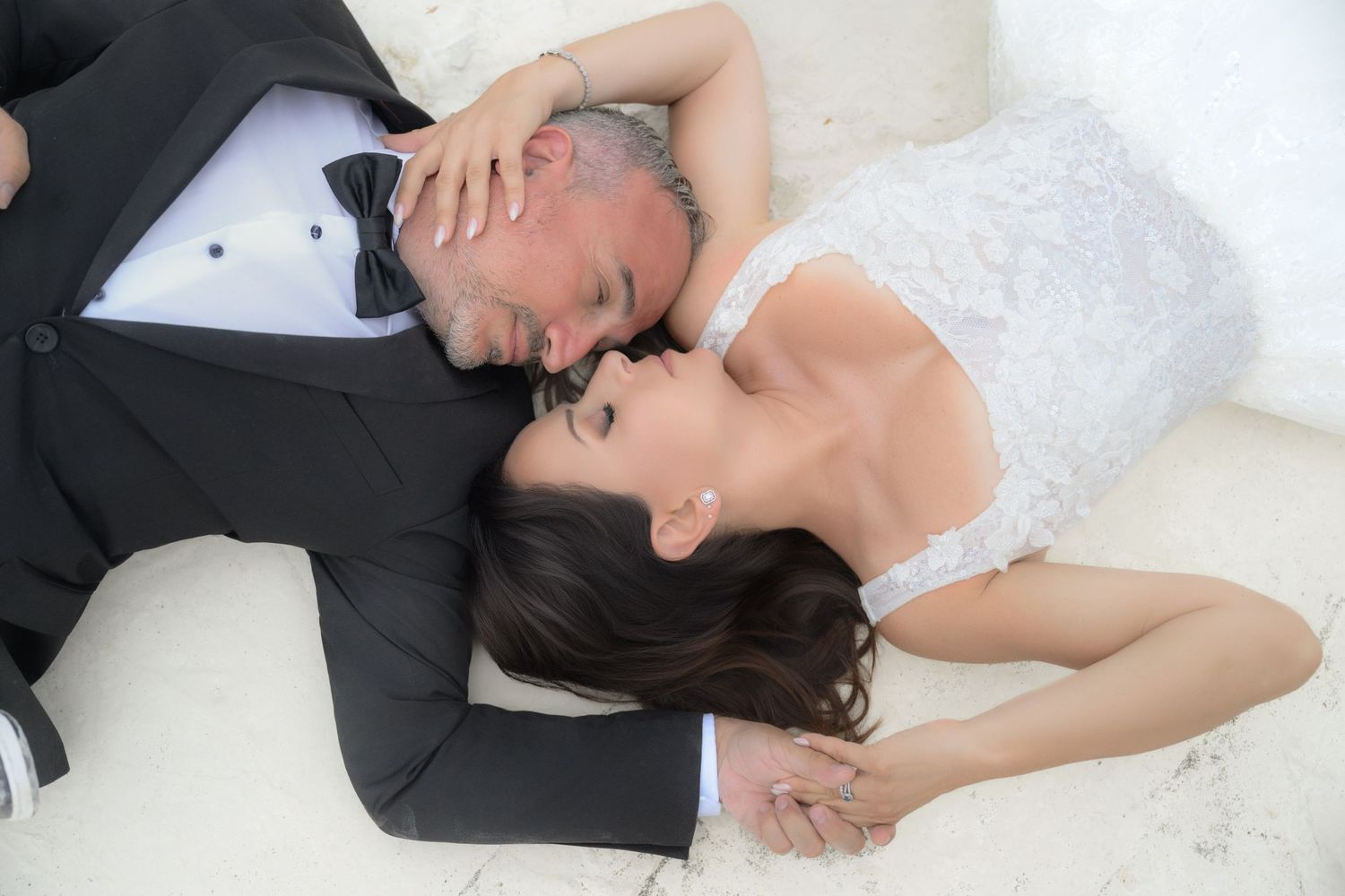 Two people in wedding attire lie close together on a white surface in an intimate pose.
