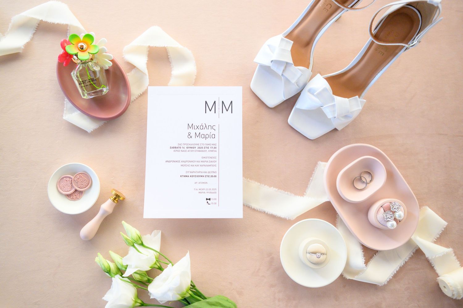 Wedding invitation with white bow sandals, coffee cups, and pink flowers arranged on beige background in flat lay style.