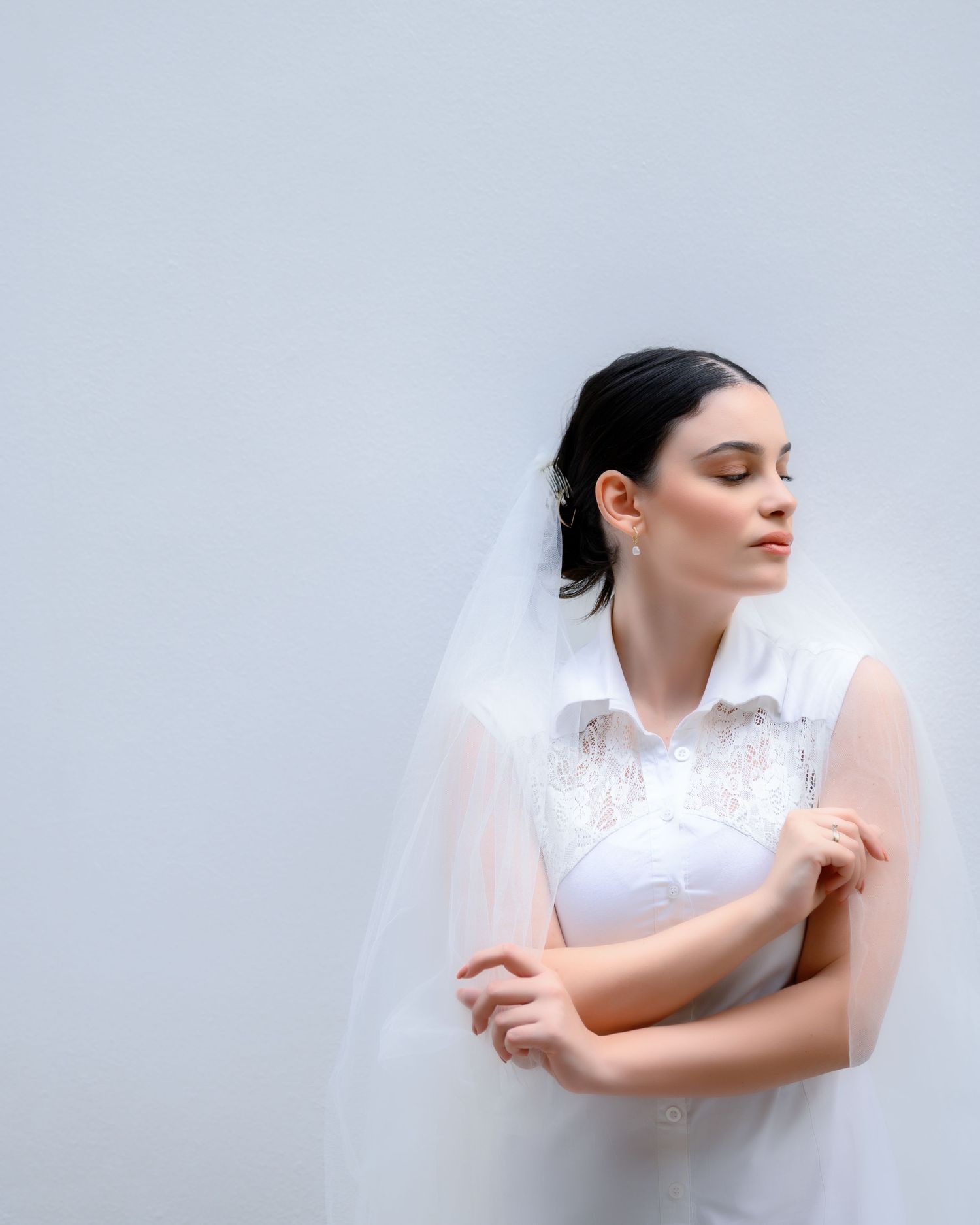 Bride in white lace wedding dress and veil stands with eyes closed against light gray background.
