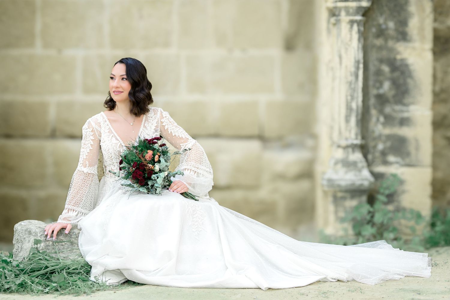 Bride in long-sleeved lace gown poses with burgundy bouquet against stone architecture.