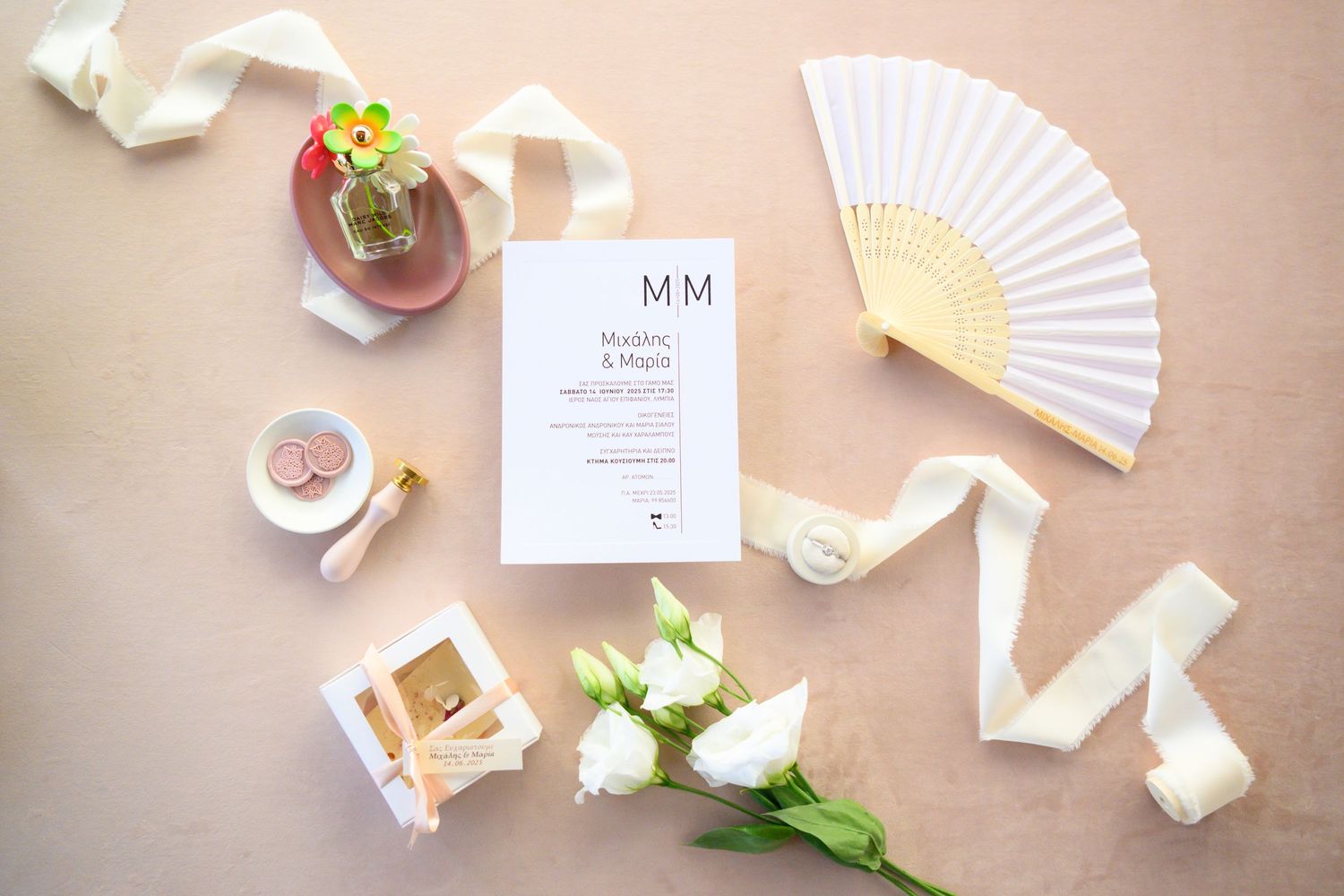 A wedding invitation suite displayed with white paper fans, pink roses, and tea cups on a blush-colored table setting.