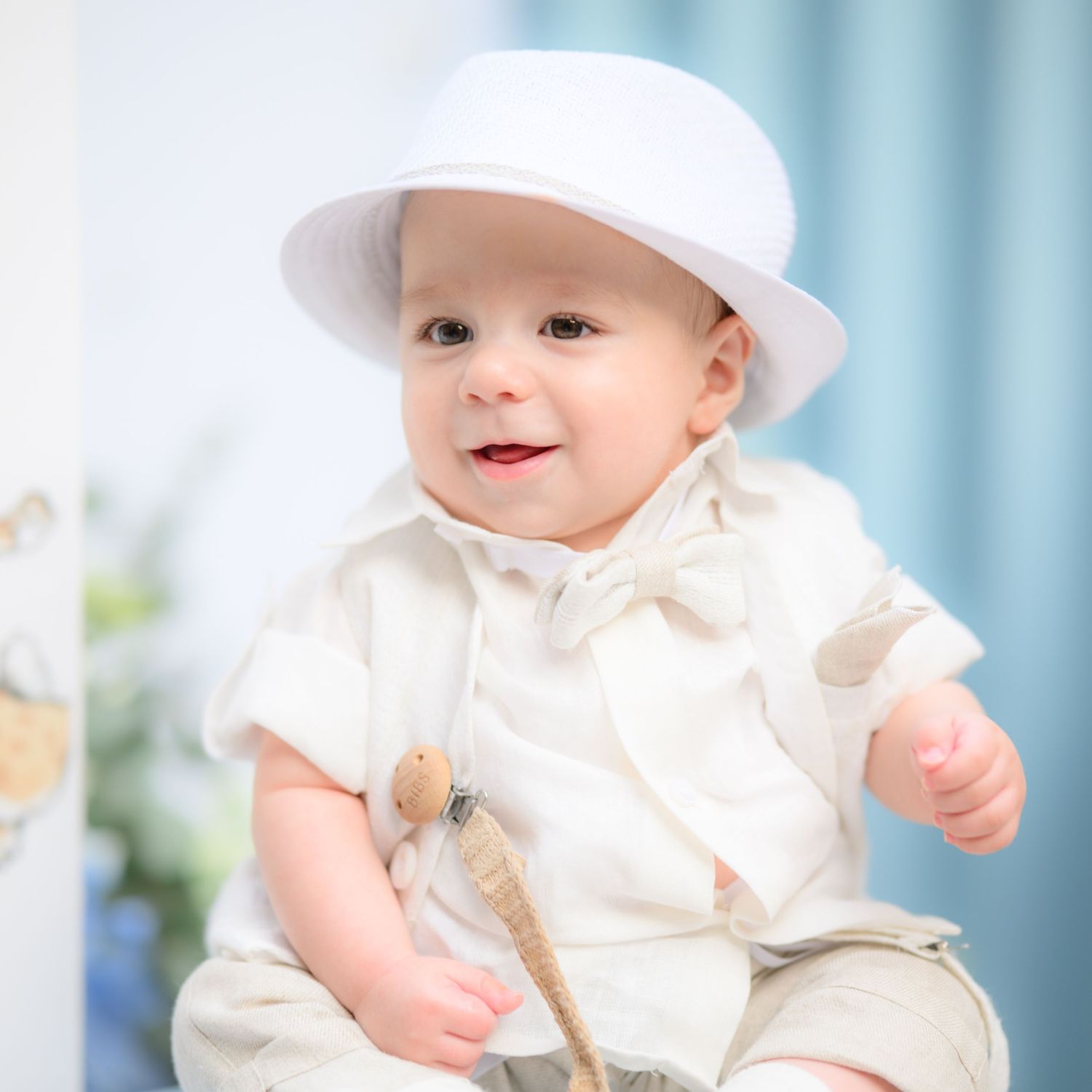 Baby dressed in white outfit and sunhat smiles while sitting against light blue background.