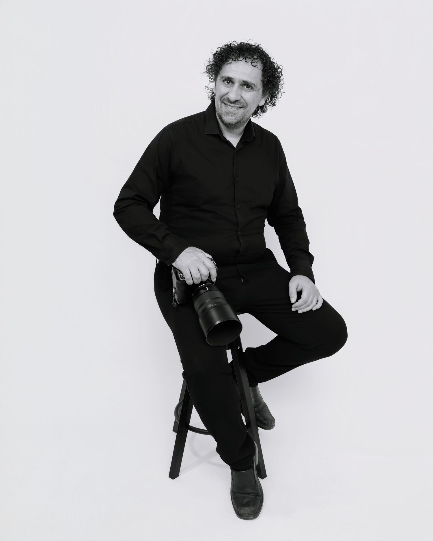 Black and white portrait of someone in dark clothing sitting casually on a stool against a white background.
