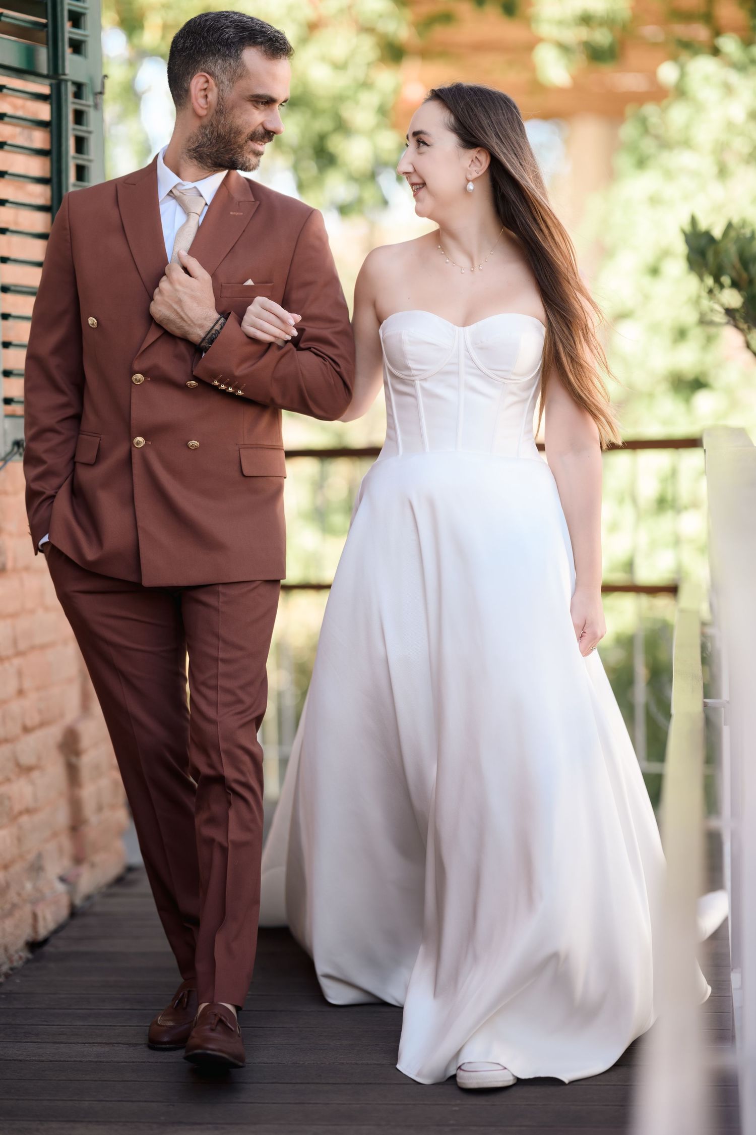 Couple in wedding attire walking together on wooden deck by brick building.