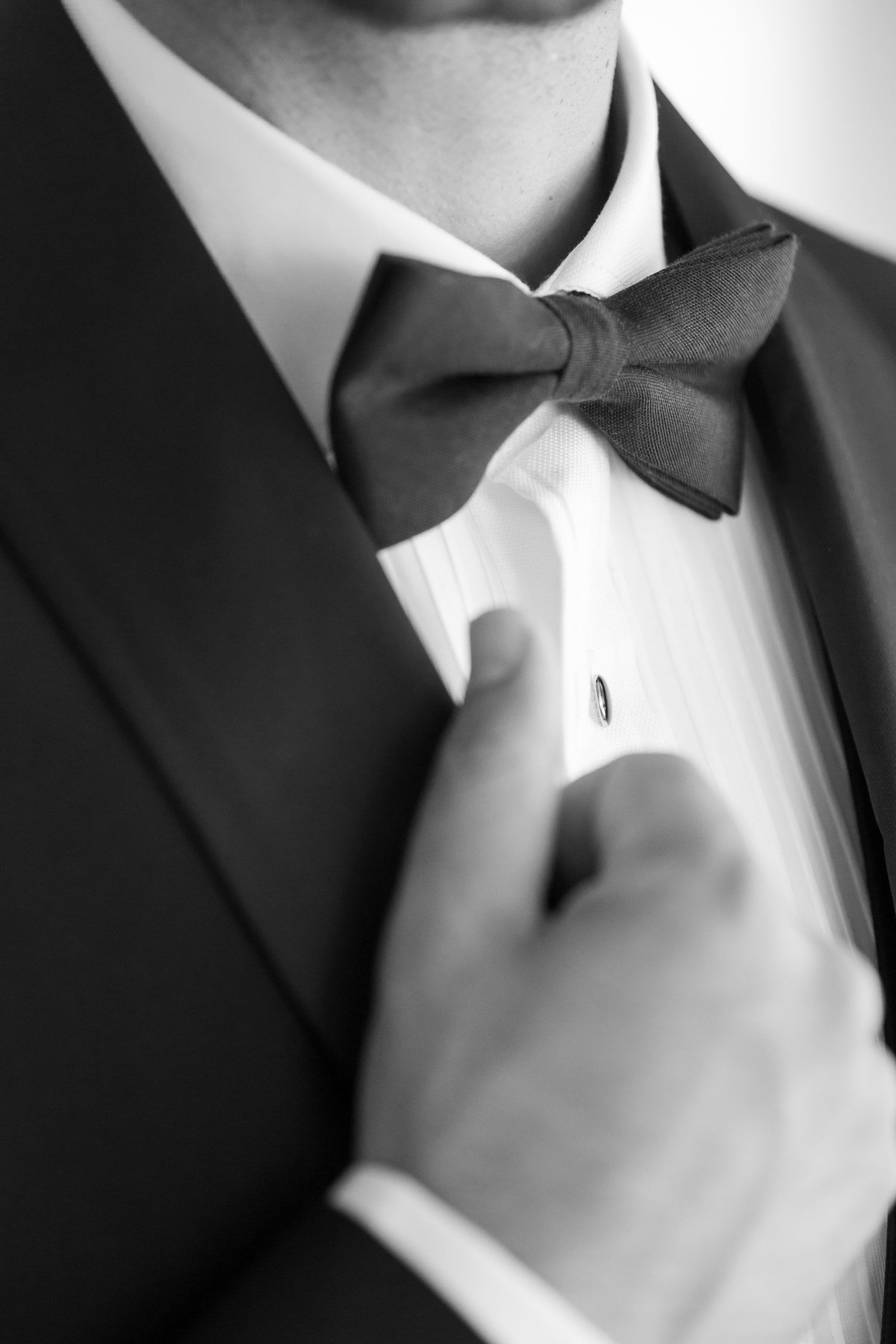 Close-up black and white photo of a black bow tie and tuxedo collar being adjusted.