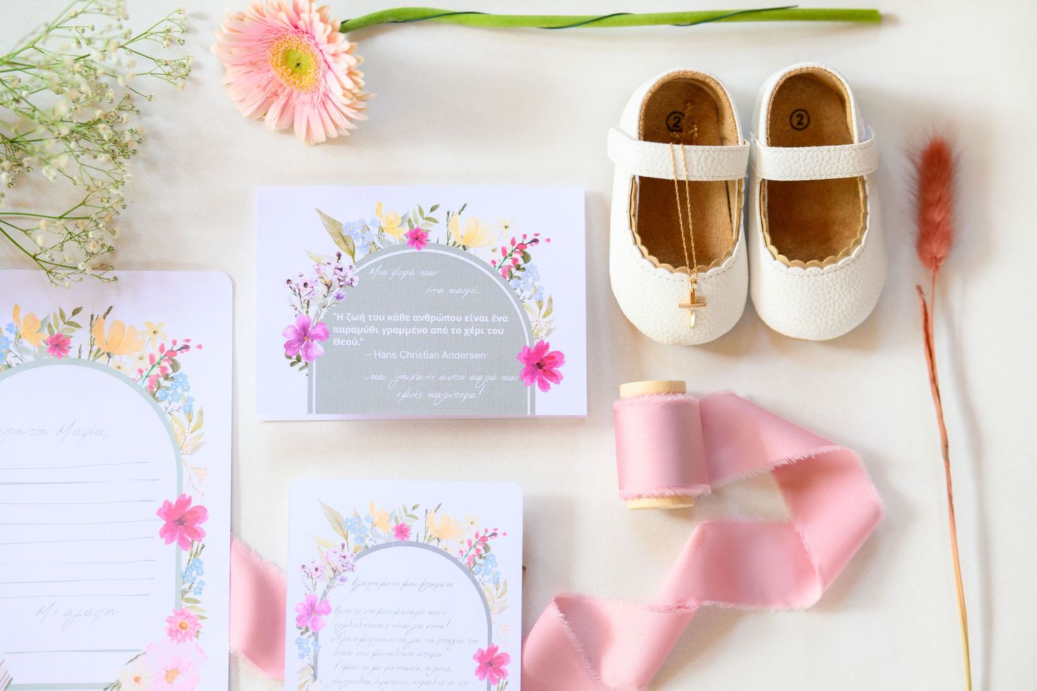White baby shoes, floral invitations, and pink ribbon arranged on a light background in a feminine baptism layout.