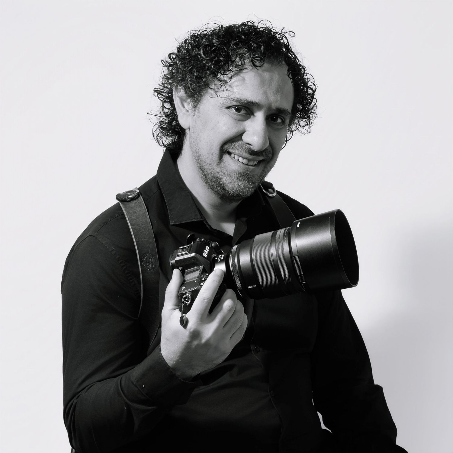 Professional photographer in black shirt smiling while holding camera with telephoto lens against white background.