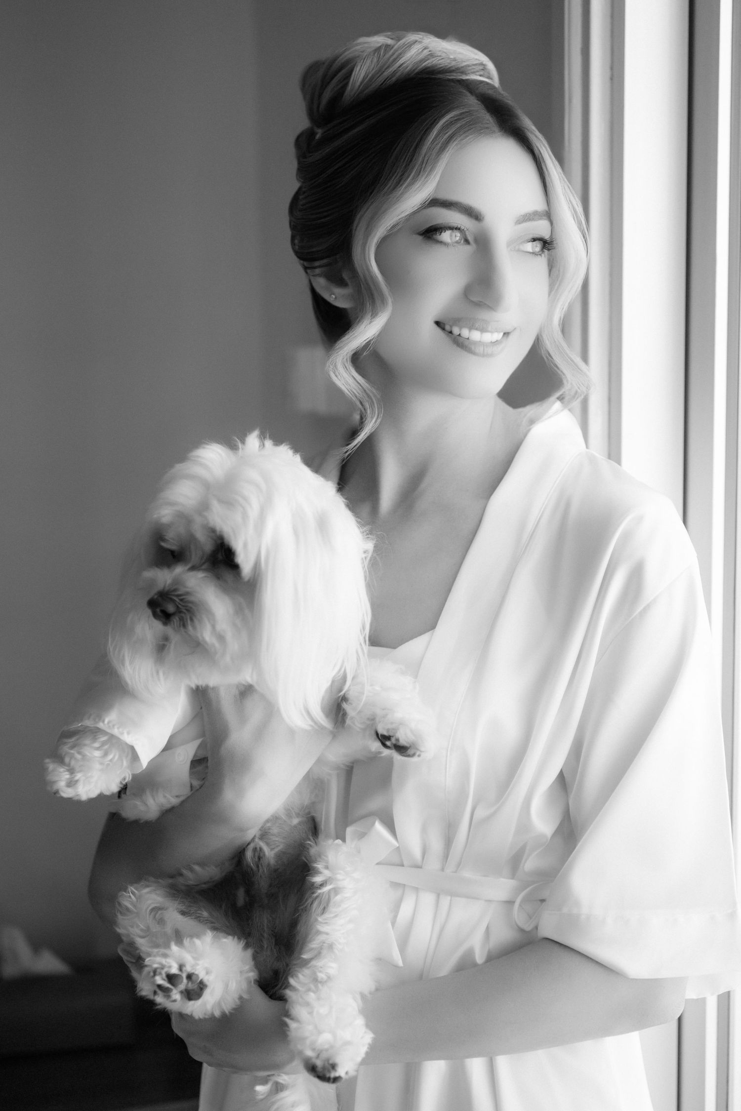 A black and white photo series shows someone in white clothing holding and playing with a small fluffy white dog near a window.