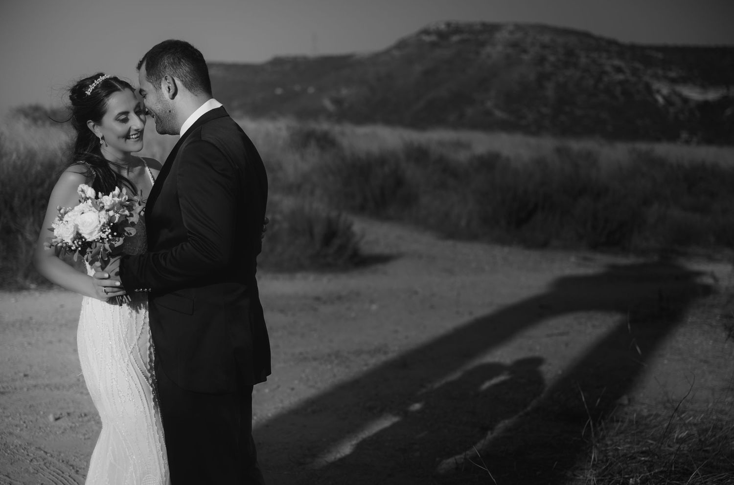 A romantic black and white wedding photo captures a sweet moment between newlyweds on a hillside at sunset.