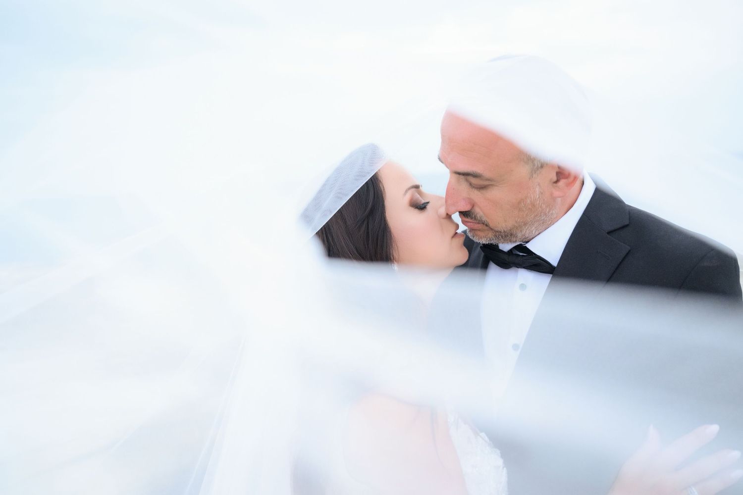Romantic bride and groom share a kiss while wrapped in a flowing white veil against a soft blue sky.