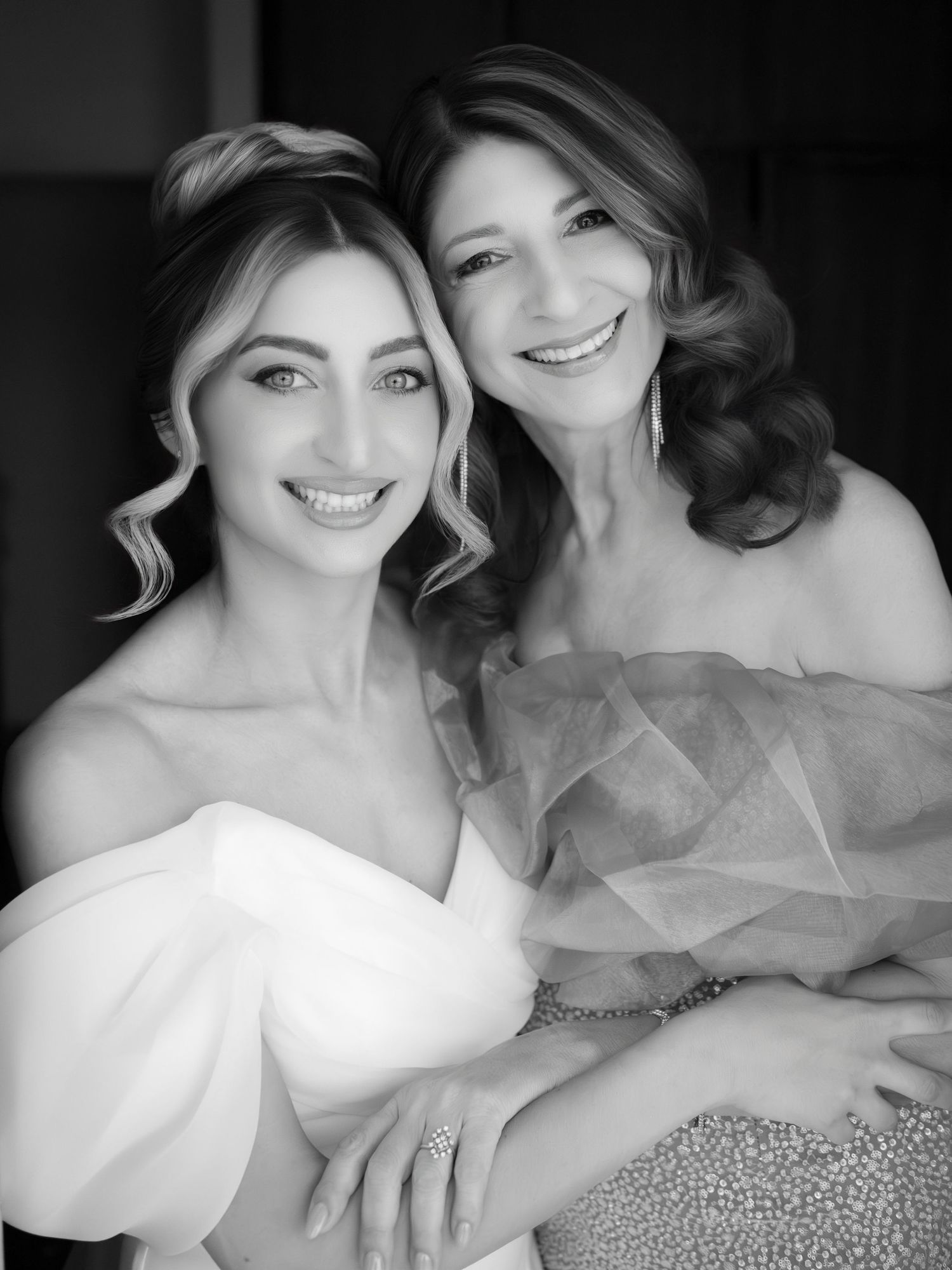 A black and white portrait captures two people in elegant white gowns with curled hair and joyful expressions.
