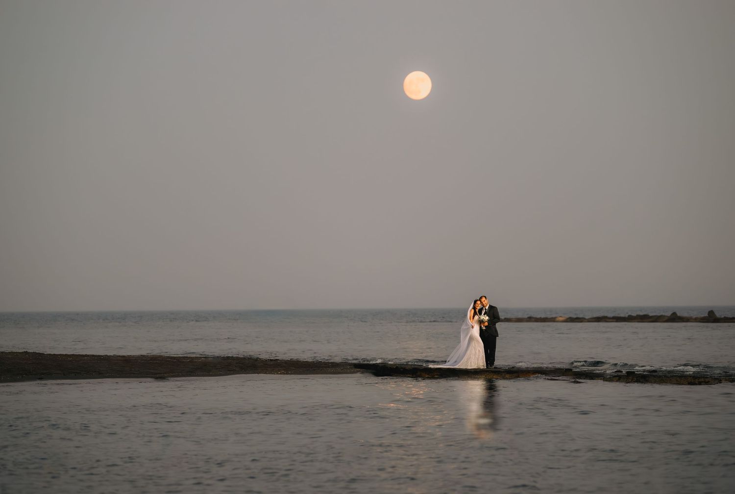 Full orange moon hanging over calm ocean waters with silhouettes of people standing on rocky coast at dusk.