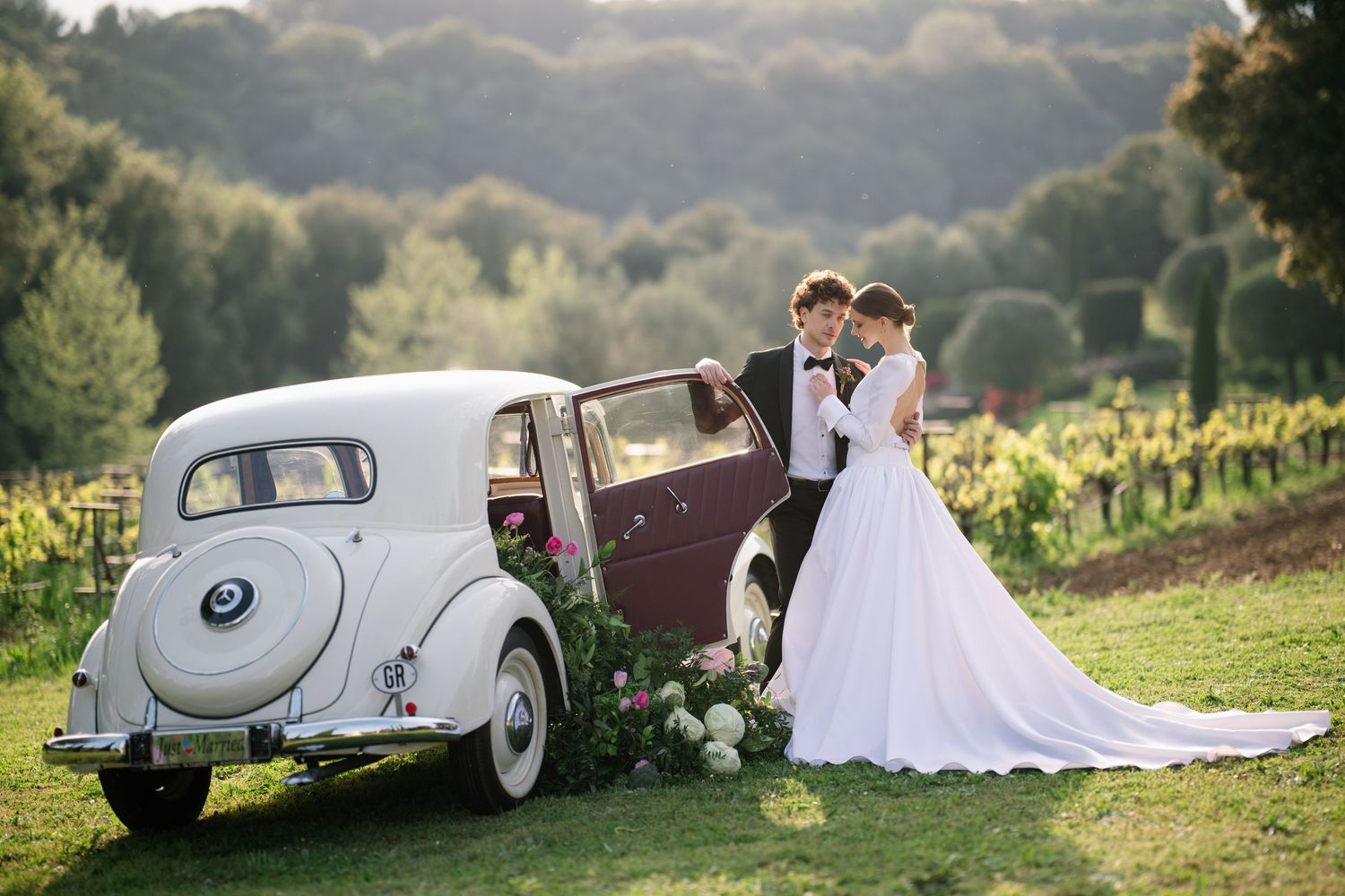 Classic vintage car parked in a scenic vineyard setting with wedding couple posing near rolling hills at sunset.