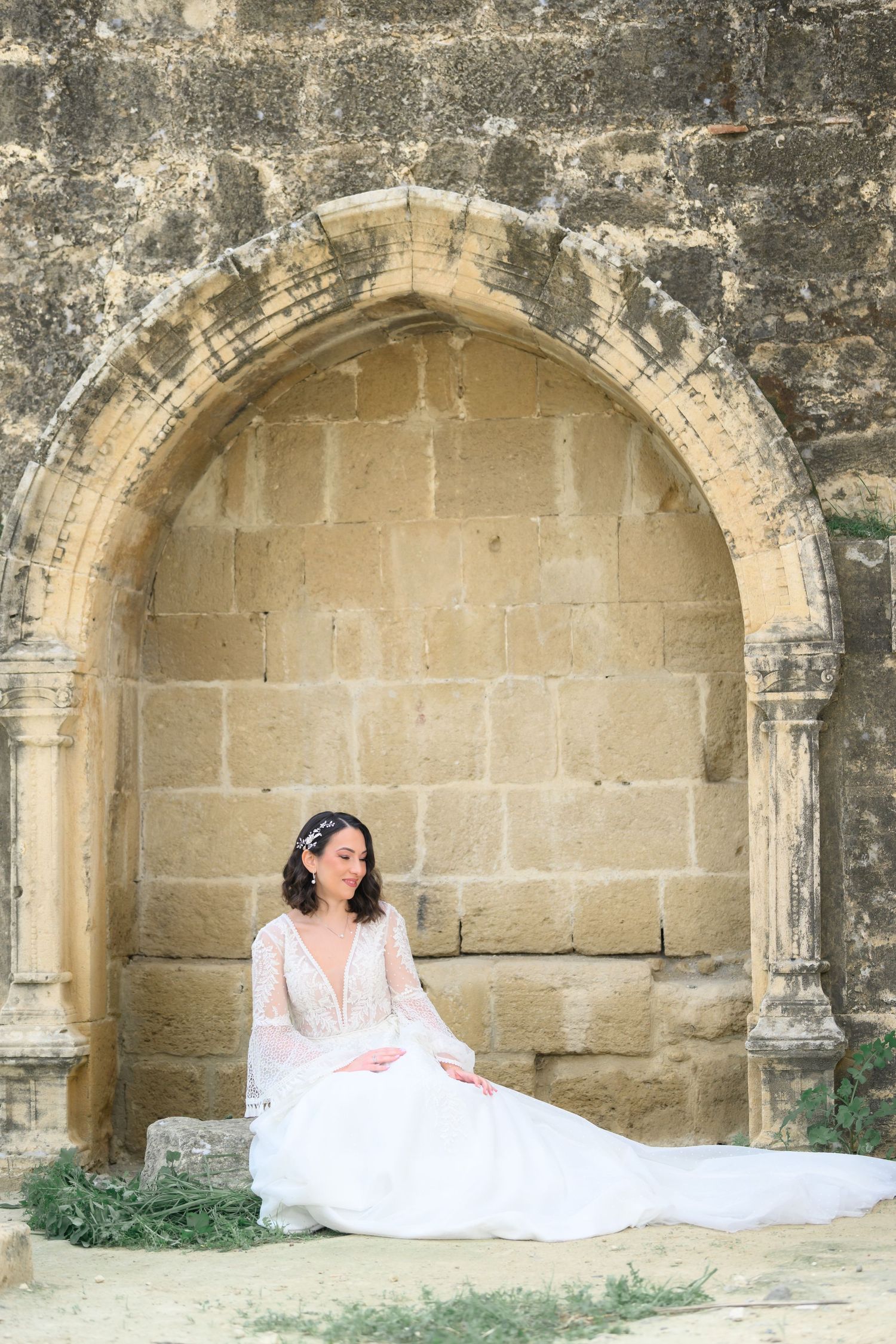 A serene stone archway alcove surrounds a person in a flowing white dress seated gracefully on stone steps.