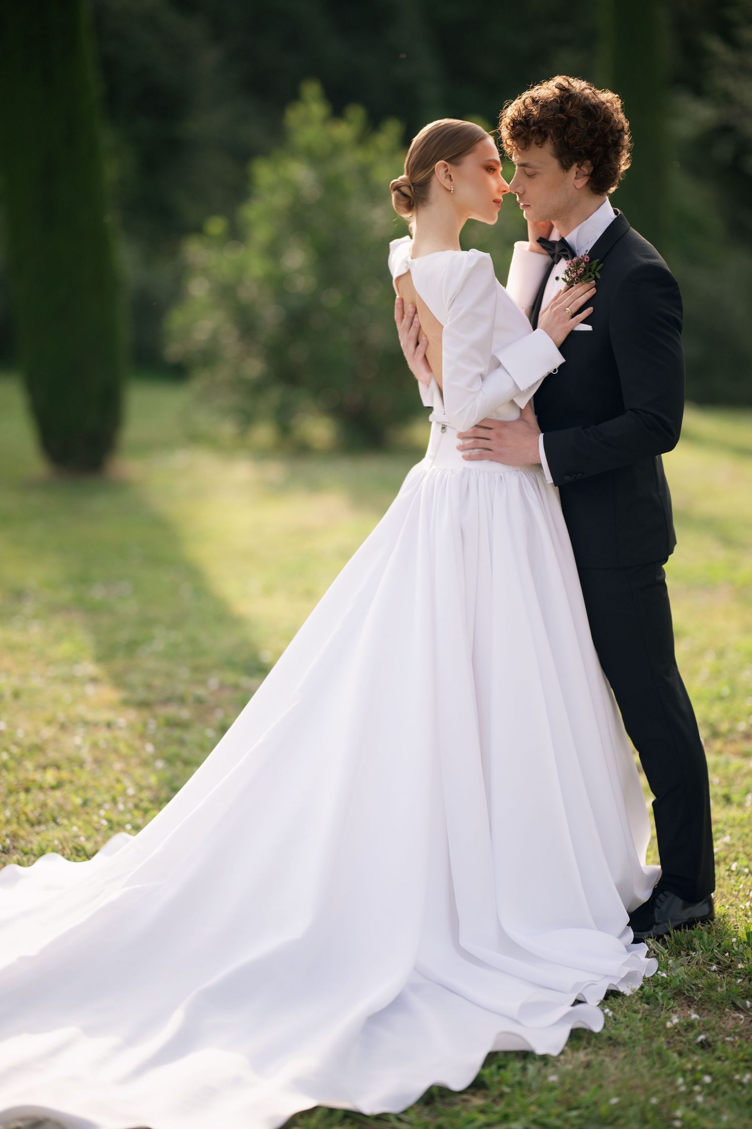 A bride in a flowing white gown and groom in black suit share romantic moments in an outdoor garden setting.