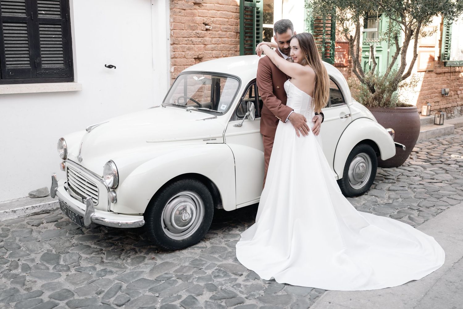 A wedding couple poses with a classic white Morris Minor car on a cobblestone street.
