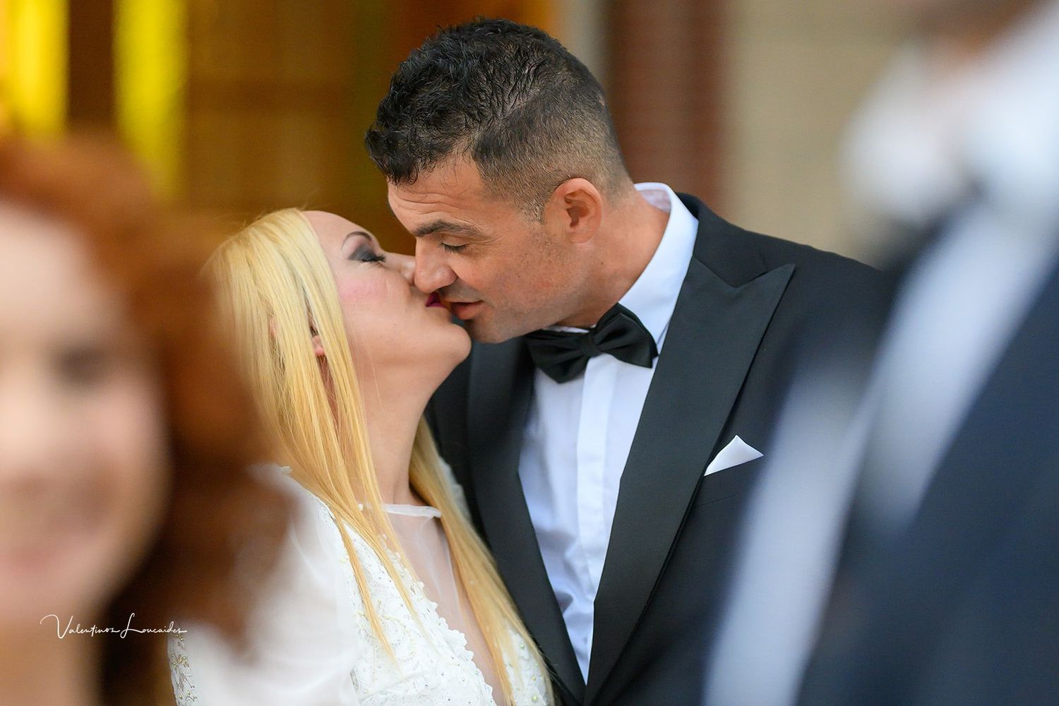 Wedding couple shares intimate kiss while guests look on in background.