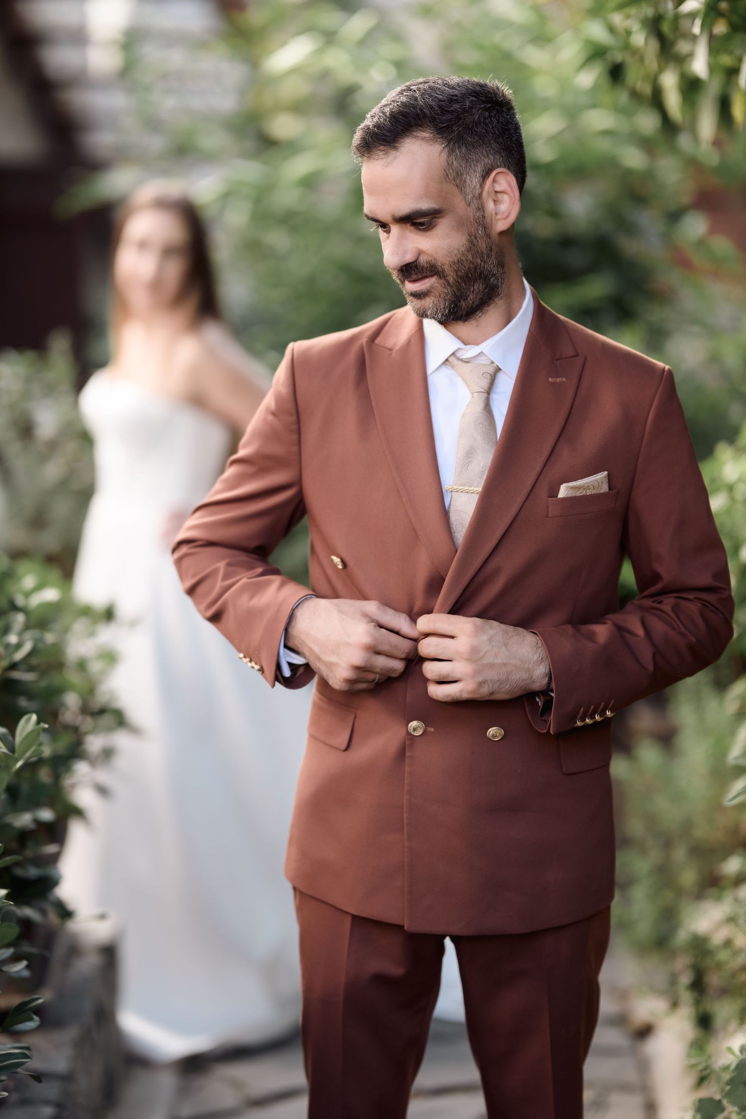Elegant groom in brown suit with white tie standing in front of bride in garden setting.