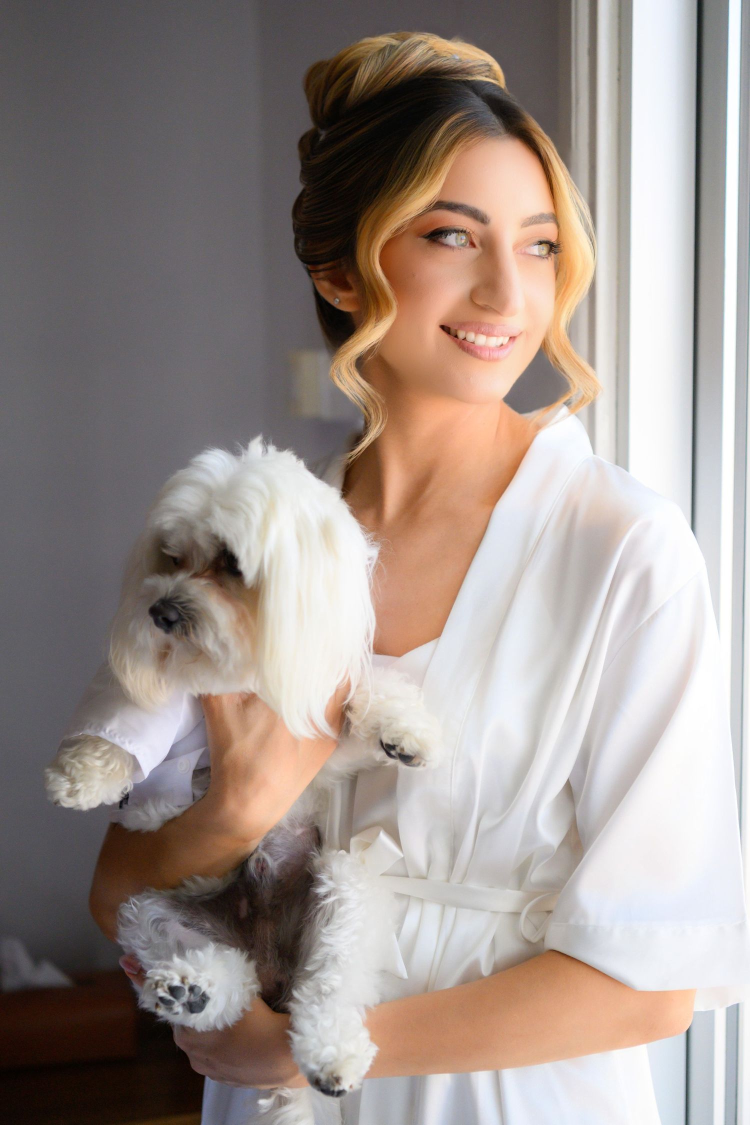 A young woman in a white robe holds a small fluffy white dog near a window in a natural light setting.