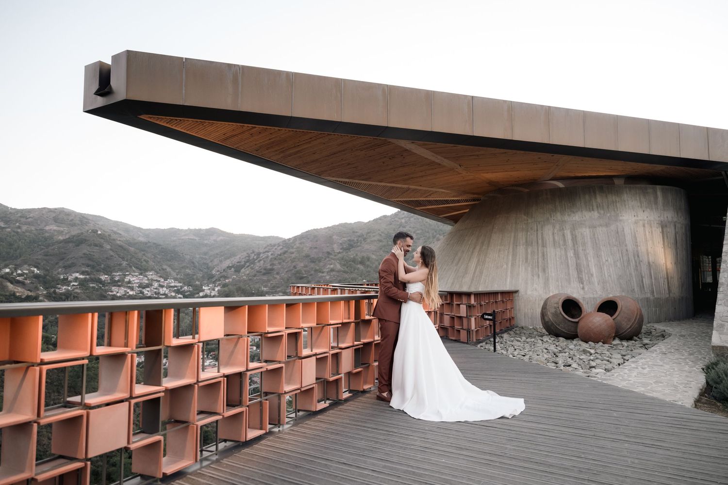 A couple stands on a modern deck with angular architecture, rust-colored metal shelving, and mountain views in the background.