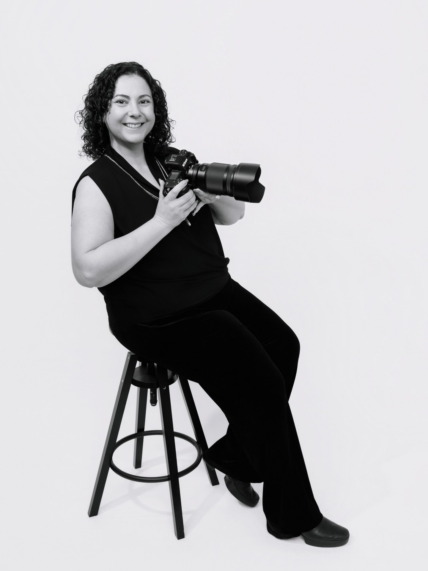 Professional photographer in black dress sits on wooden stool with camera in creative studio portrait series.