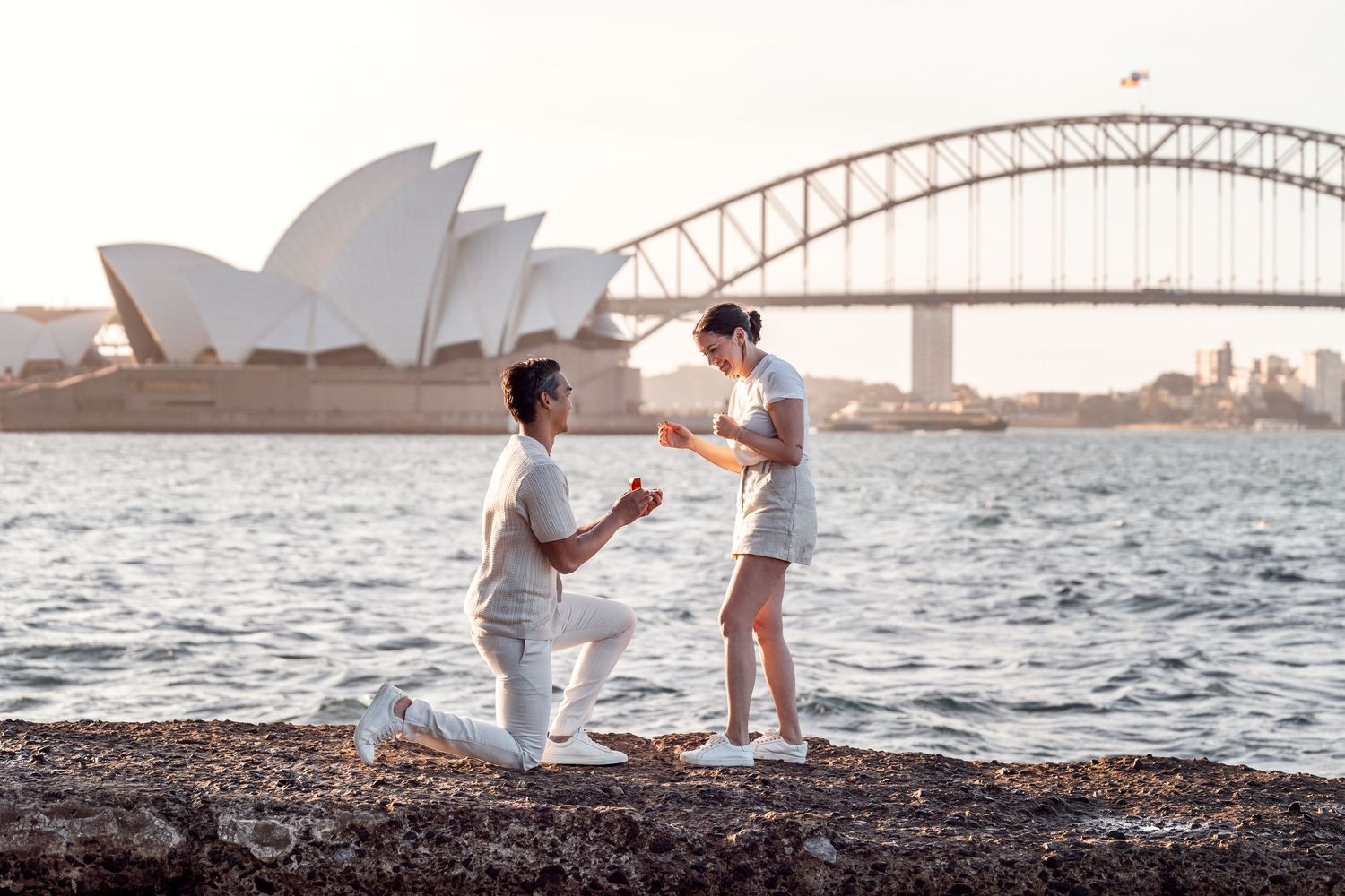 a man is kneeling down in front of the harbour bridge and the opera house asking a girl to merry him during a secret proposal photoshoot