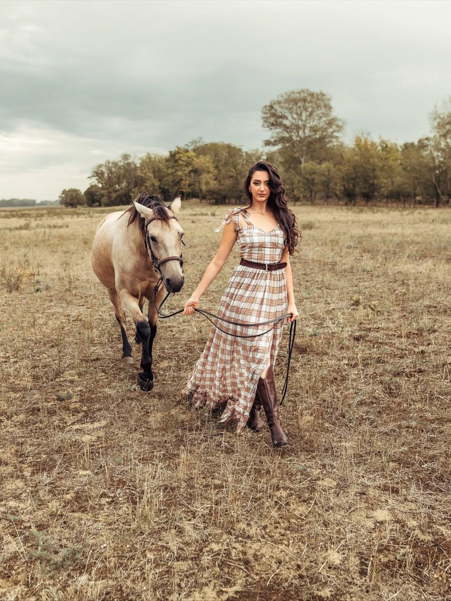 A person in a plaid dress walks with a light-colored horse across a dry autumn field at sunset during a photoshoot in sydney