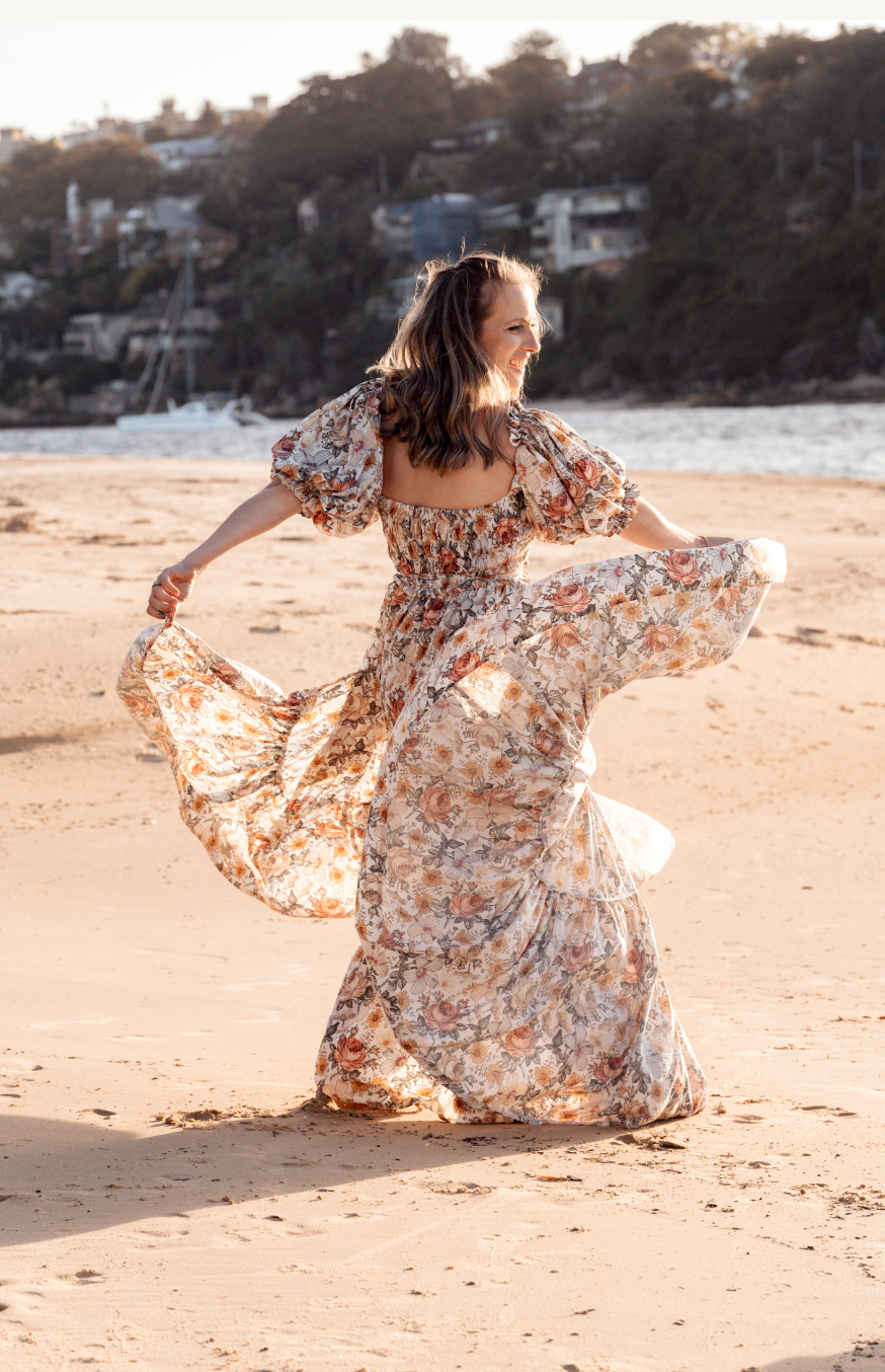 A flowy floral dress billows gracefully as someone dances and twirls on a sandy beach at sunset.