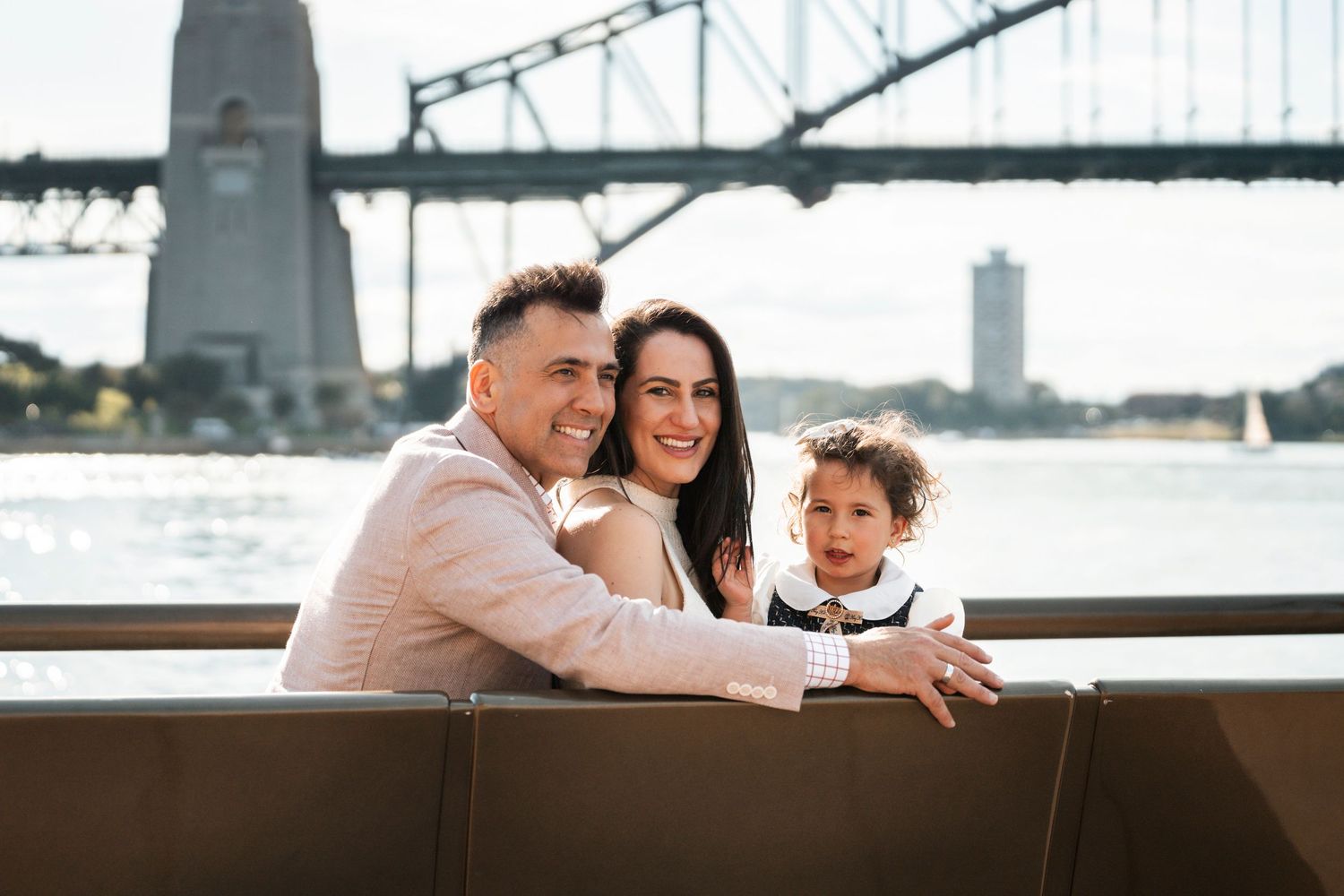 A family enjoys a sunny day together by the Sydney Harbour Bridge, smiling and leaning on a bench during a family photoshoot in CBD Sydney